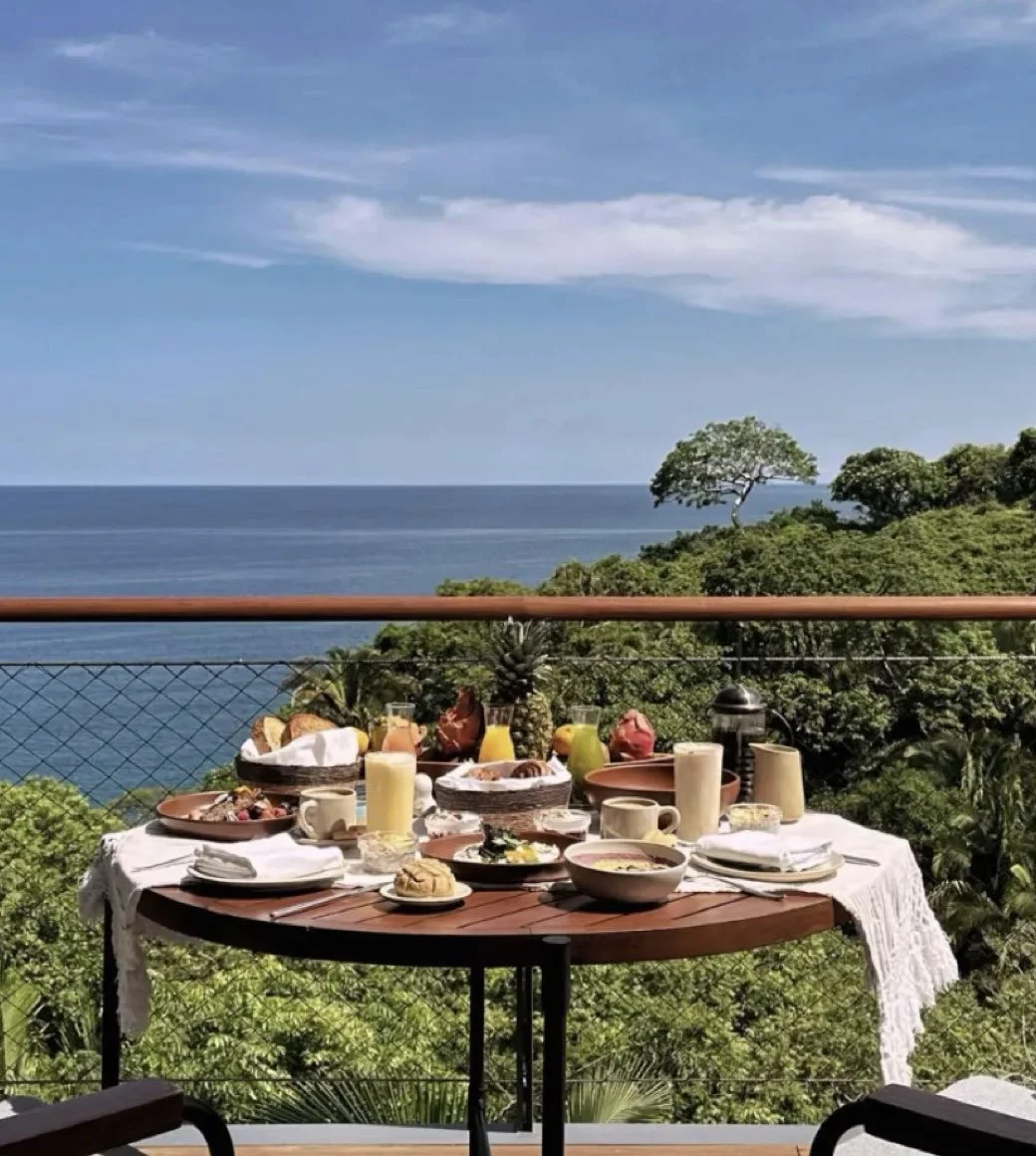 A breakfast table set outdoors on a balcony with a view of lush green trees, ocean, and partly cloudy sky. The table has various dishes, fruits, drinks, and tableware on a white cloth.
