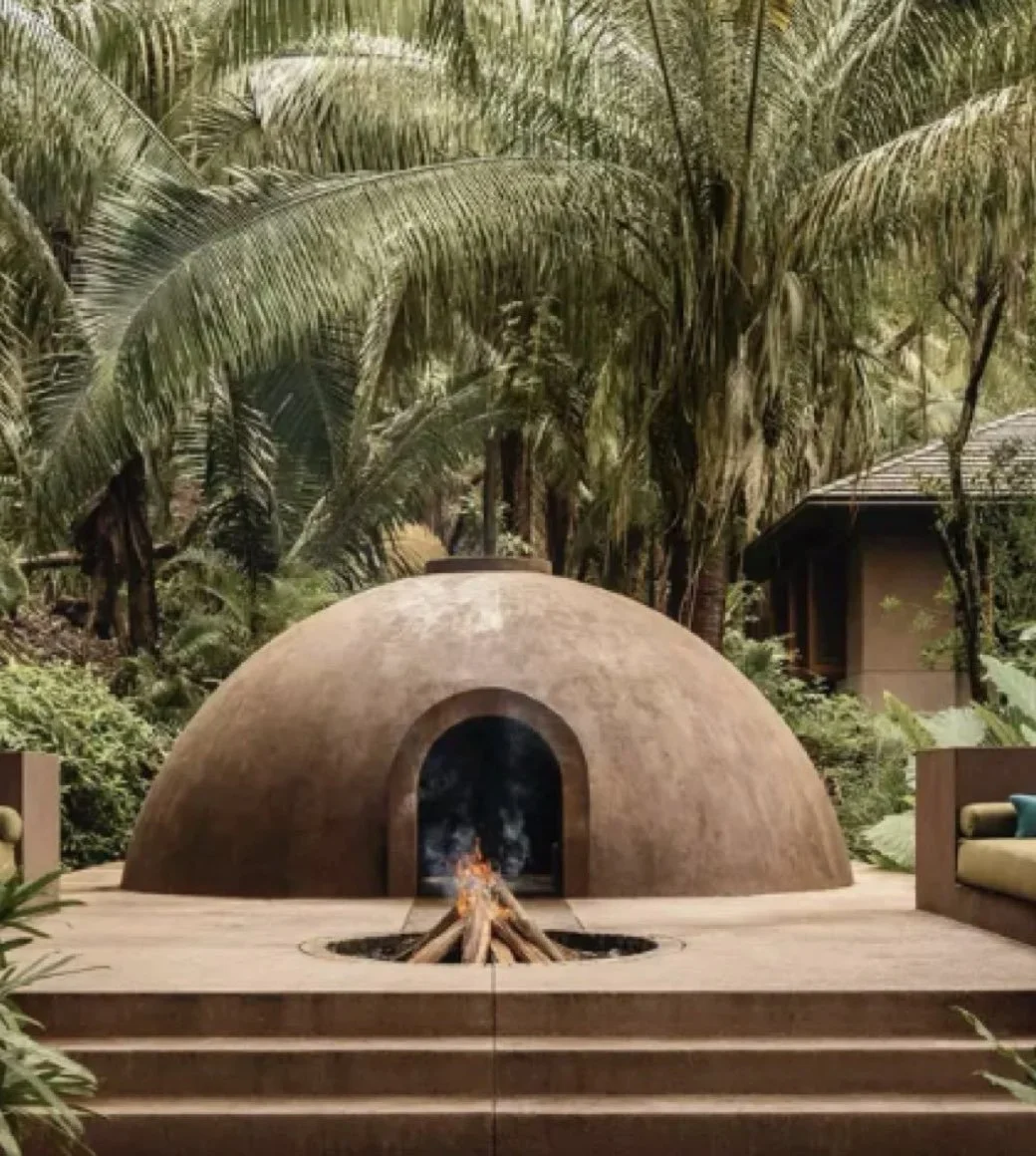 A dome-shaped outdoor fireplace with a small fire inside, set on a wooden deck surrounded by tropical plants and trees.
