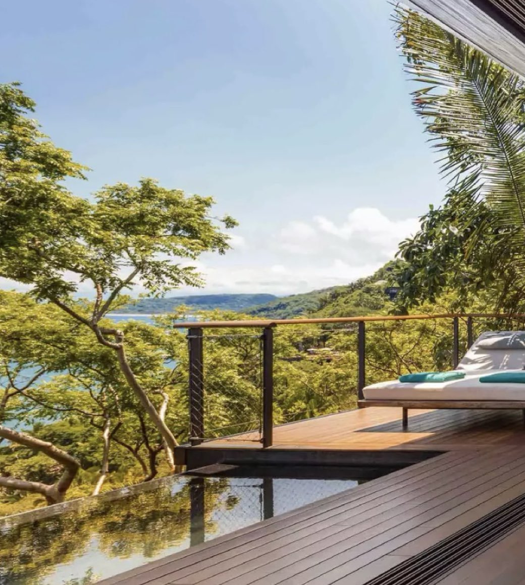 Balcony with lounge chair and towel overlooking lush green trees and a body of water under a partly cloudy sky.