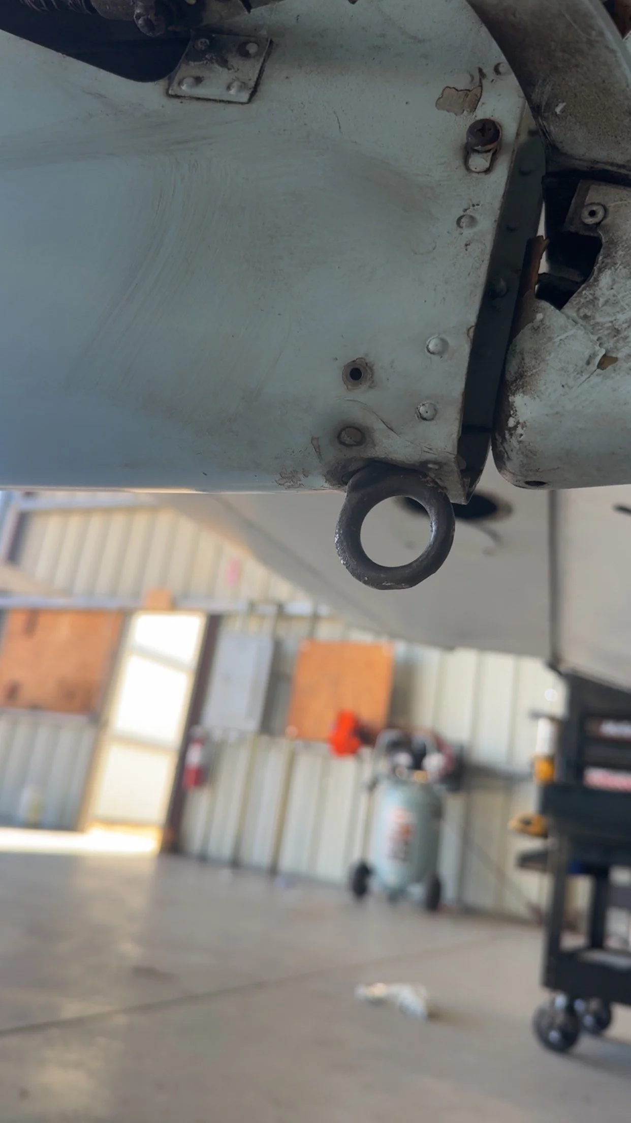 Close-up of a metal part with a black eyelet, possibly part of a boat or machinery, in a workshop or garage with tools and equipment in the background.