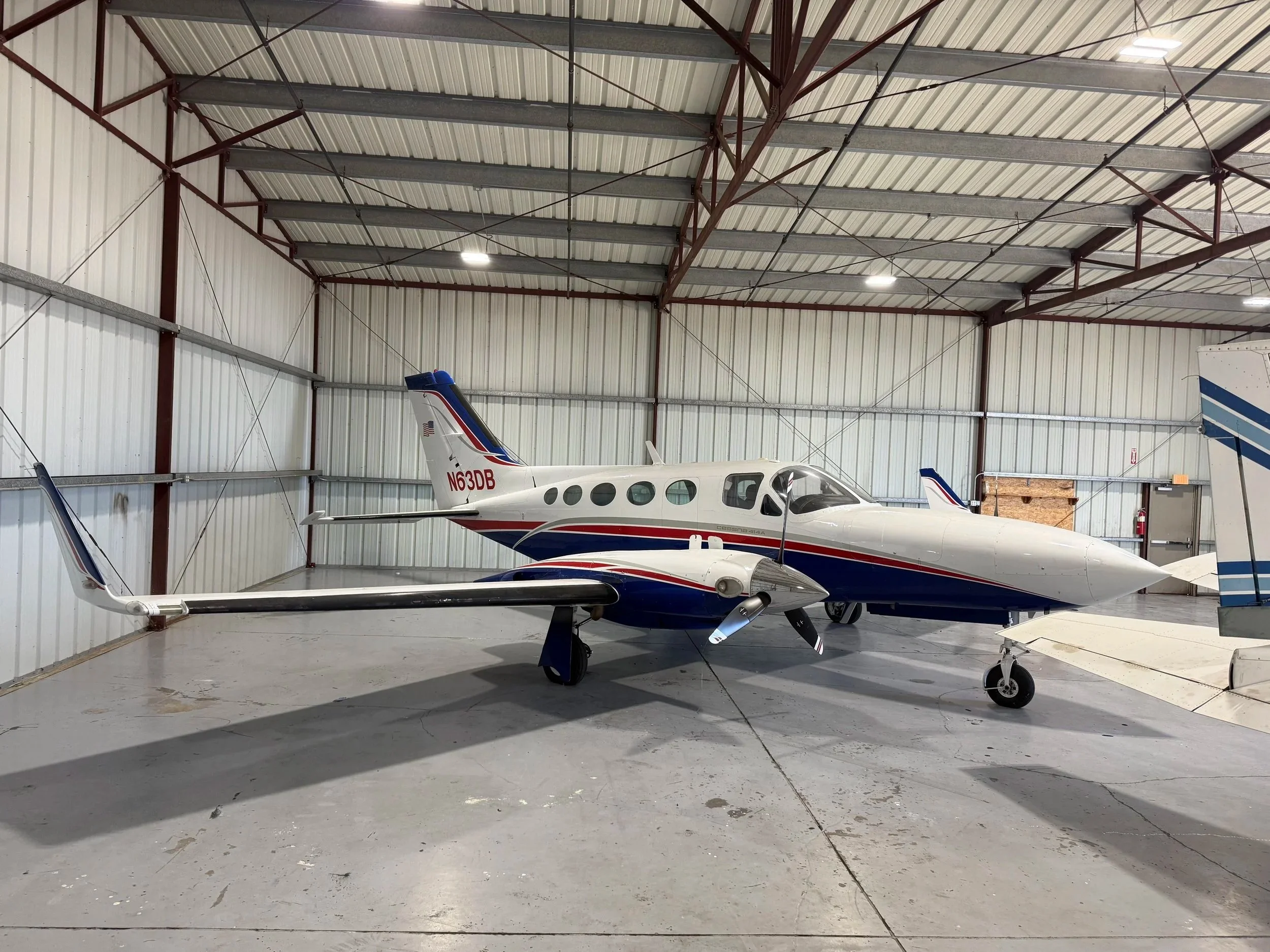 A small private jet with a white fuselage, blue and red accents, and the registration N63DB parked inside a hangar with metal walls and ceiling.