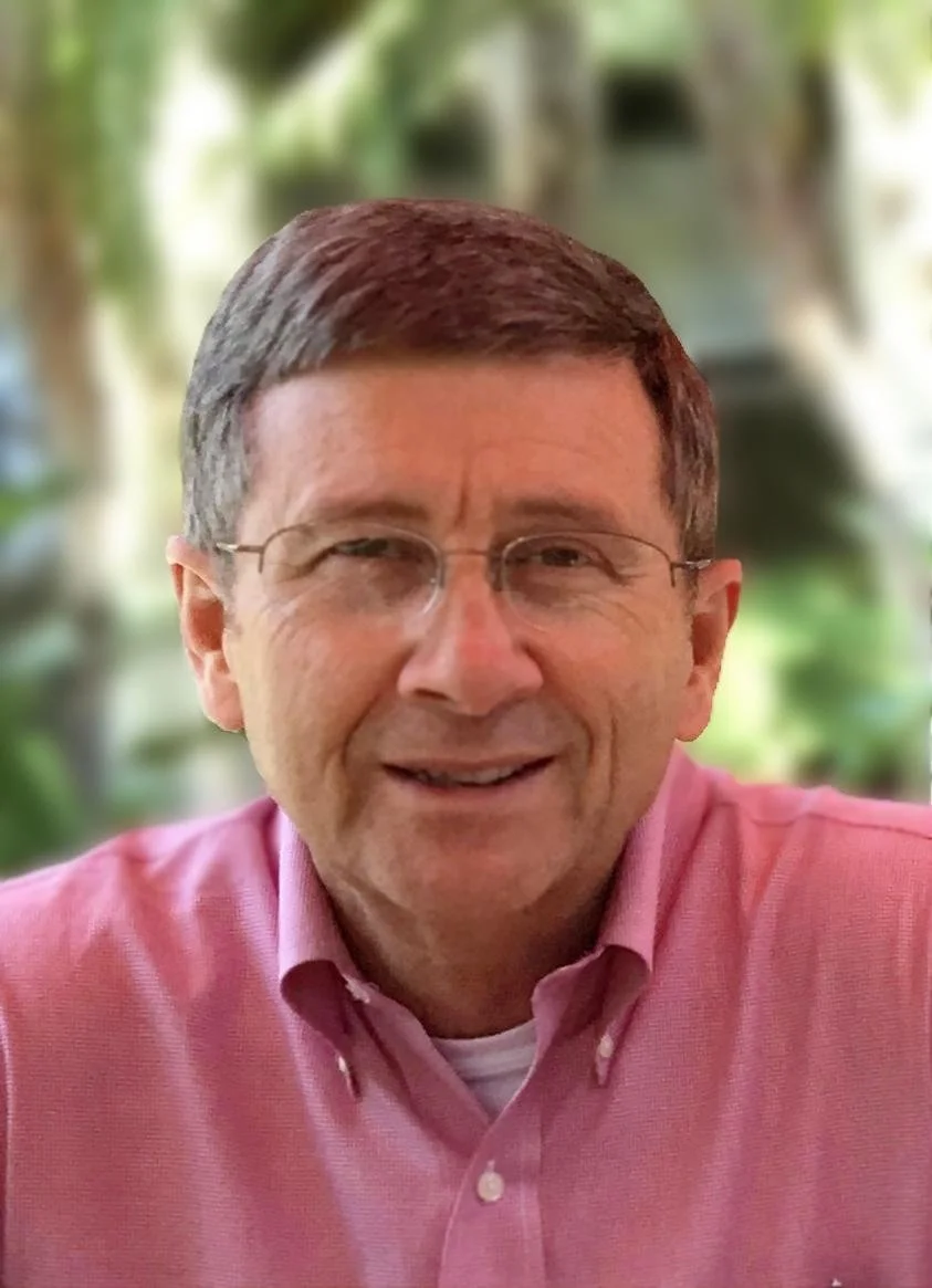 A man wearing glasses and a pink button-up shirt smiling at the camera, with a blurry green outdoor background.