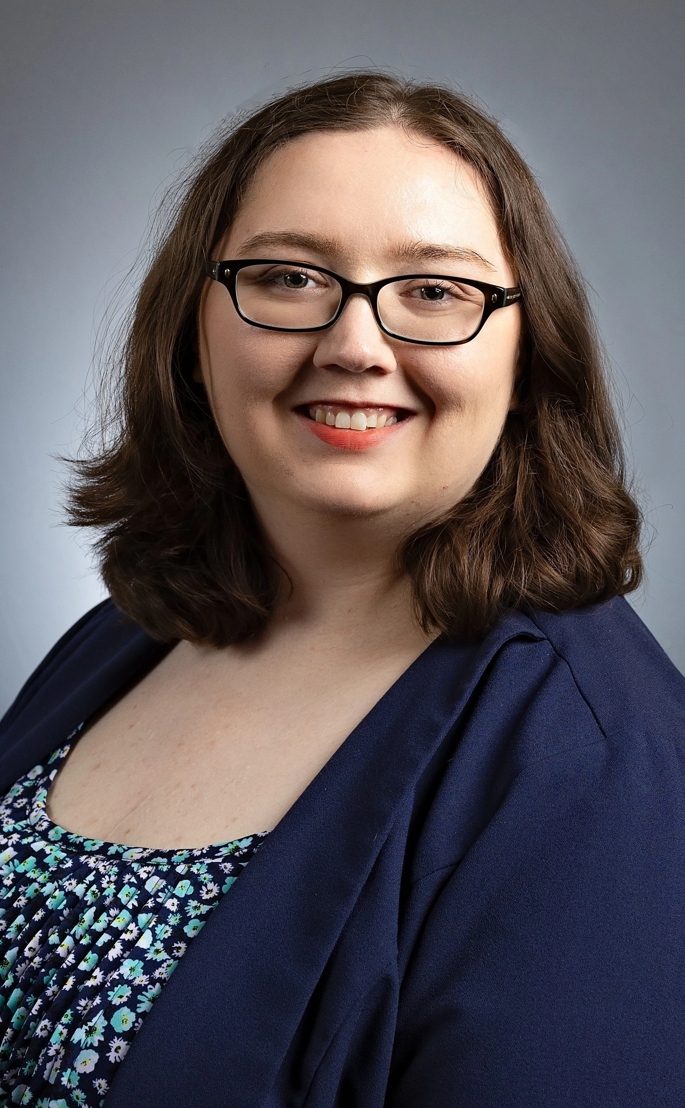 A woman with shoulder-length brown hair, glasses, and a friendly smile wearing a navy blazer and a floral top against a gray background.