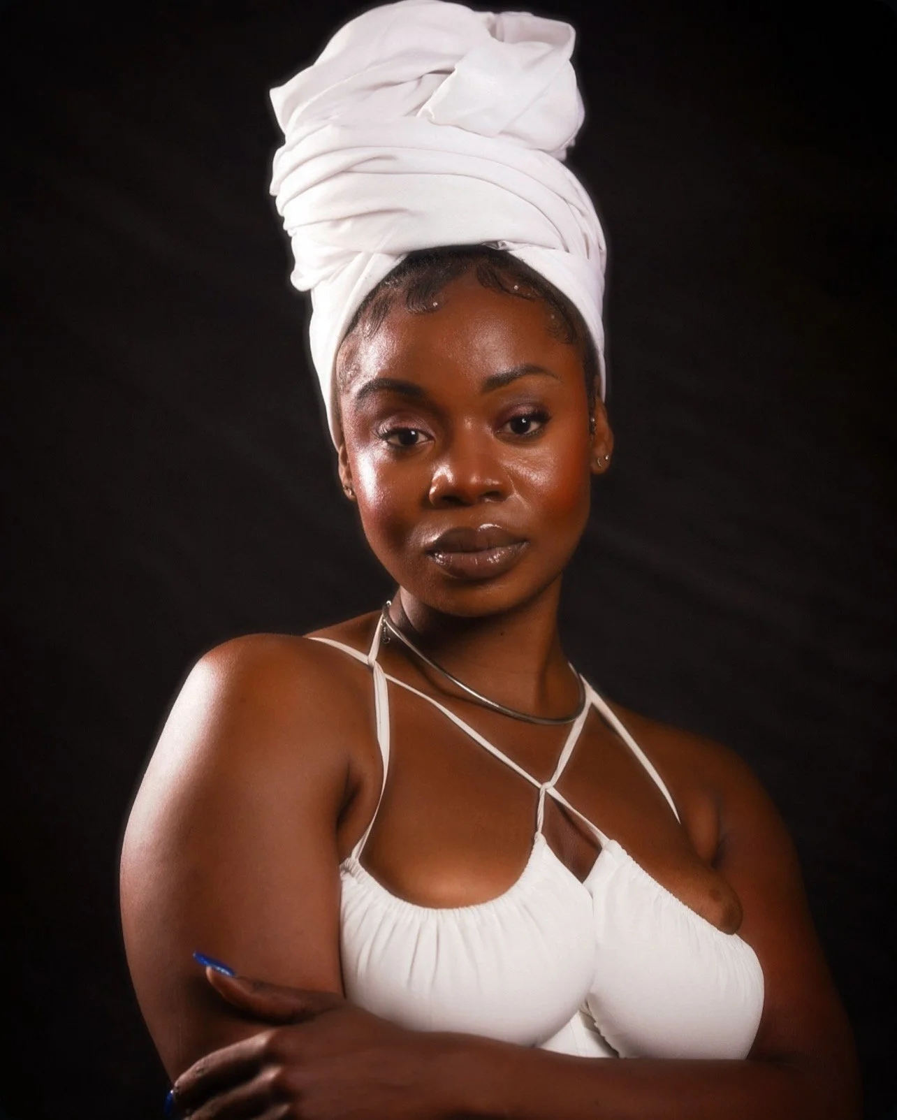 A young woman with dark skin, wearing a white head wrap and a white top with thin straps, standing against a black background.