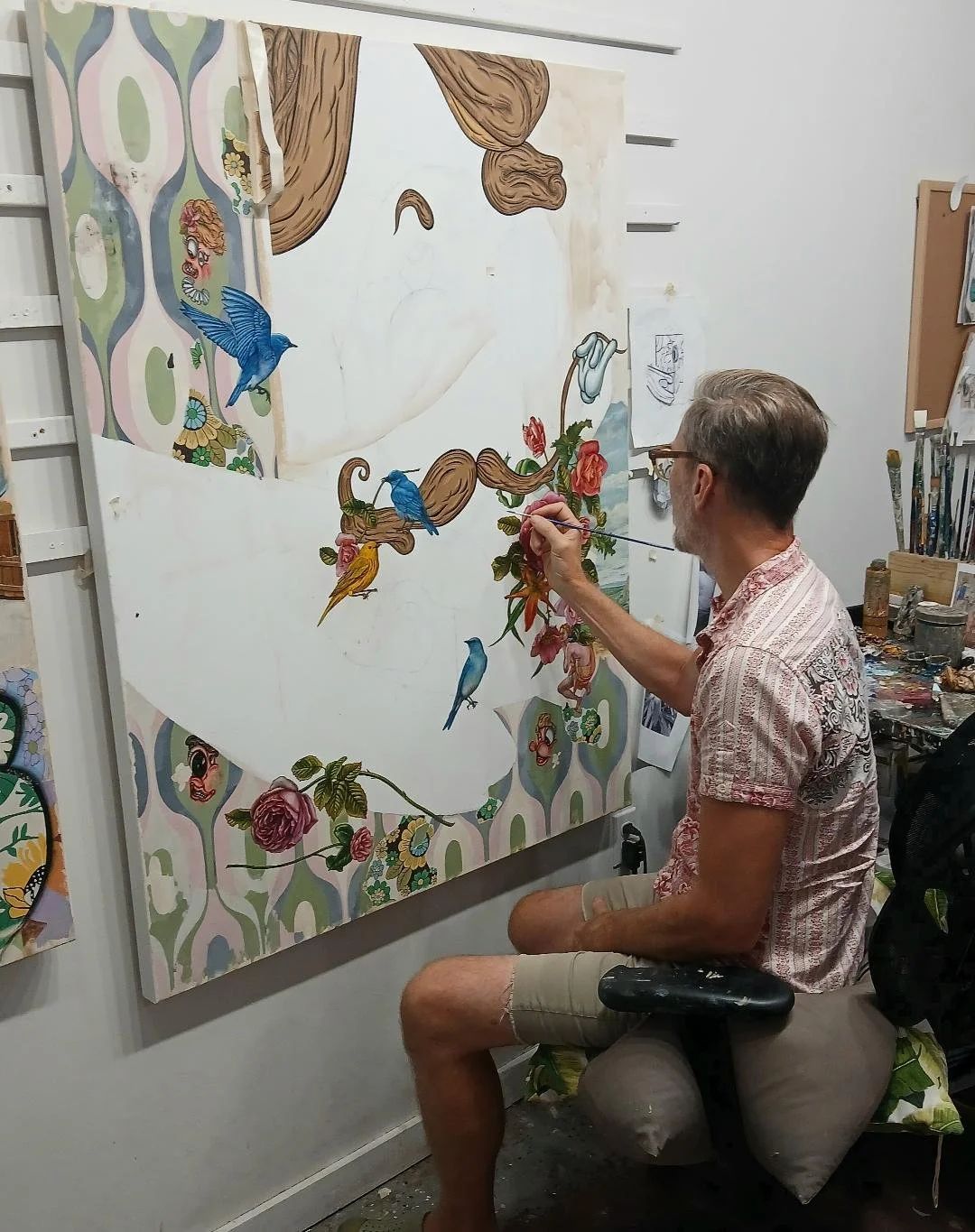 An artist painting a colorful, detailed mural on a large canvas in a studio, with various art supplies and sketches visible in the background.