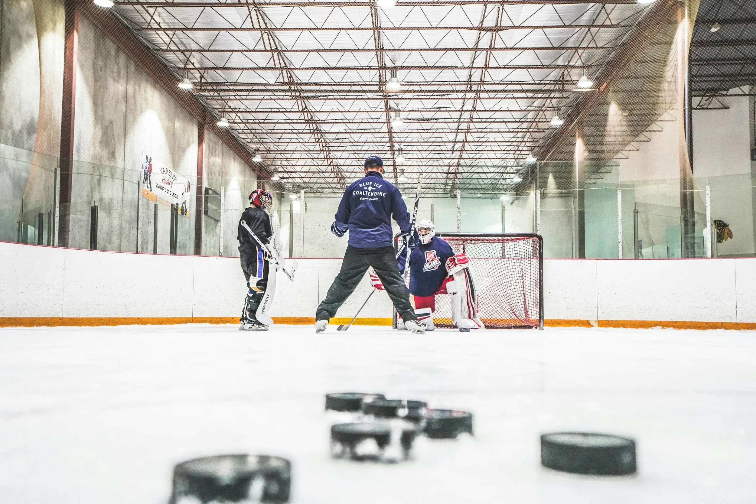Hockey training session at an indoor rink with two young players, a coach, and a goalie practicing near the net, with hockey pucks on the ice in the foreground.