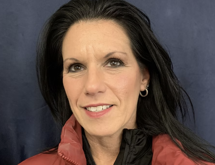 A woman with long black hair and hoop earrings smiling at the camera, wearing a red jacket against a dark background.