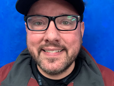 A man wearing glasses, a black cap, and a black and red jacket, smiling in front of a blue background.