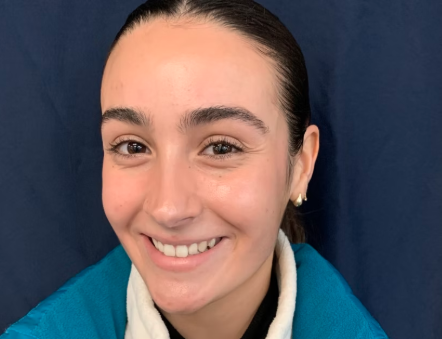 Close-up of a smiling young woman with dark hair pulled back, wearing a blue jacket and pearl earrings, against a dark background.