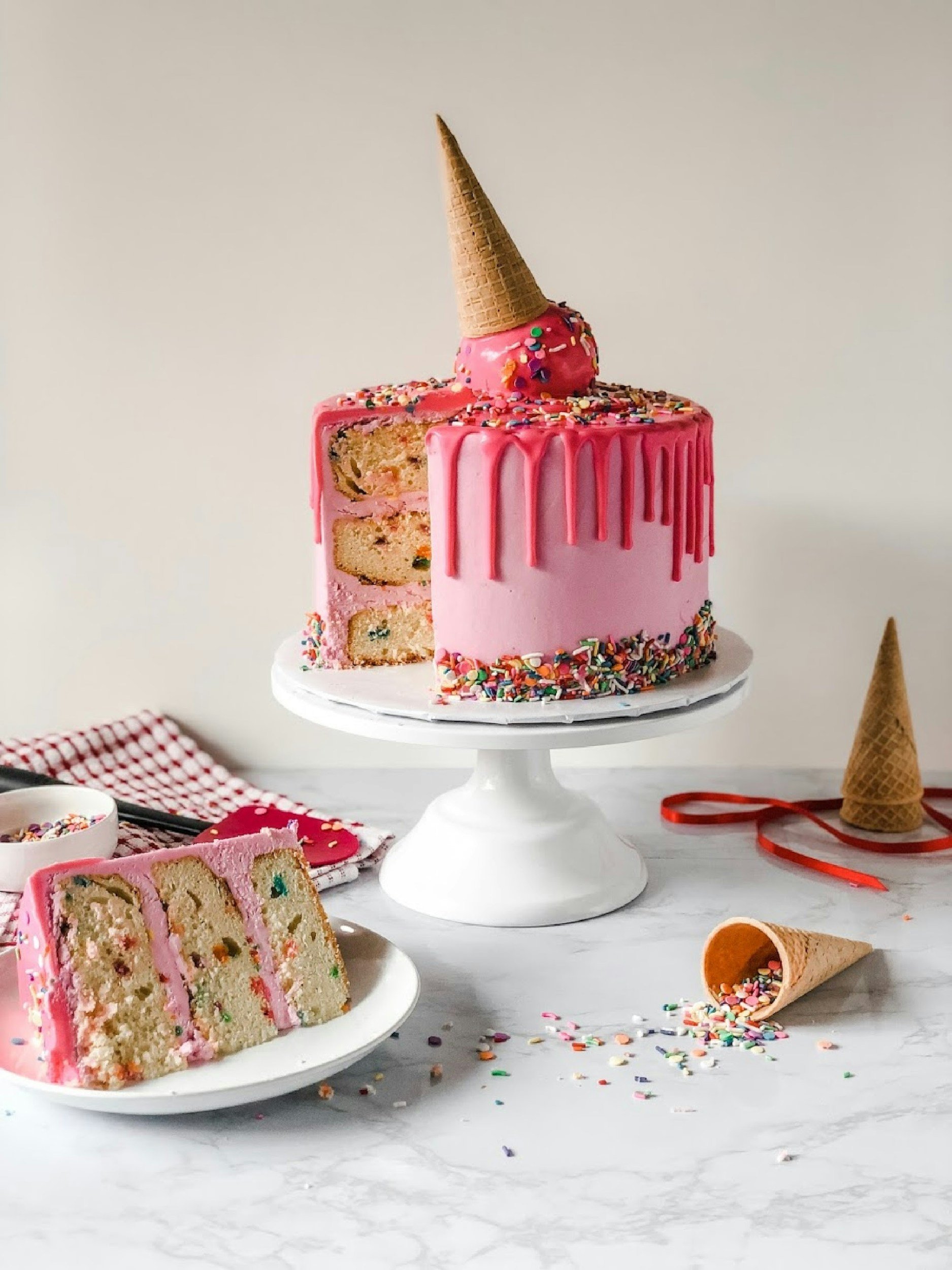 Pink layered cake with rainbow sprinkles, pink drip icing, an upside-down ice cream cone on top, and a slice cut out showing colorful interior. Two ice cream cones and spilled sprinkles on a marble surface.