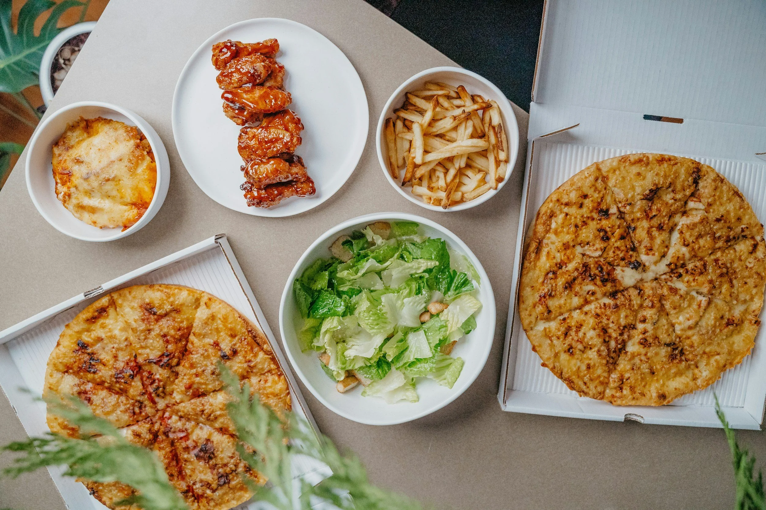 Two large pizzas in open boxes, one with pepperoni and the other with cheese and toppings, along with side dishes including a green salad, French fries, chicken wings, baked cheese dip, and a small bowl of cheese topping.