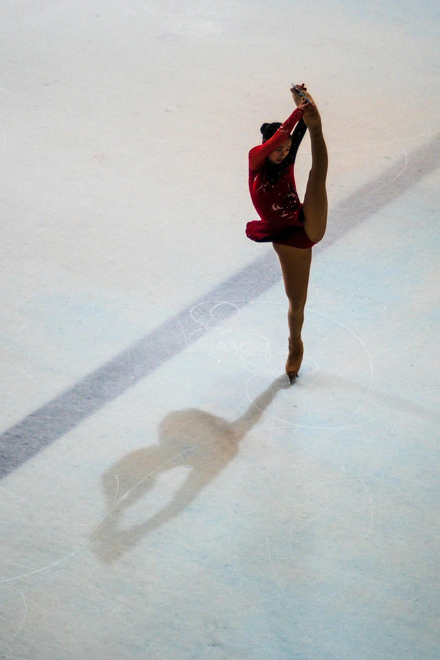 A female figure skater in a red dress performing a high-kick on the ice rink, creating a shadow on the ice.