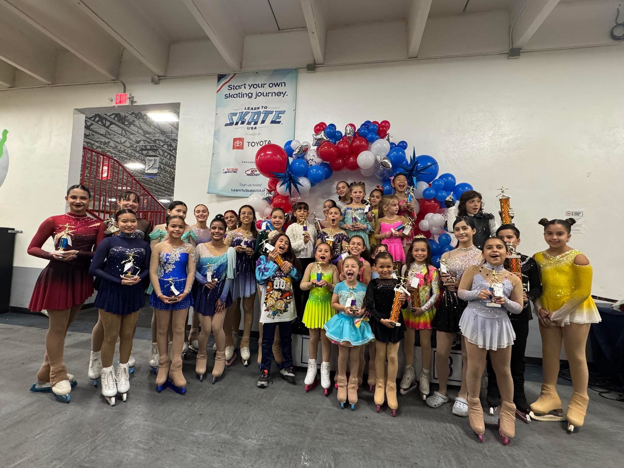 Group of young female figure skaters in colorful costumes holding trophies, celebrating their competition with a decorated backdrop of balloons and a banner that reads 'Learn to Skate USA'.