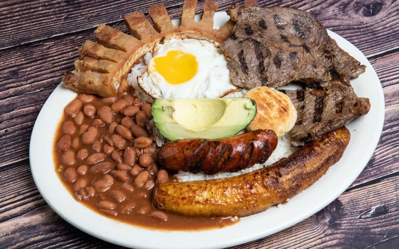 Plate of traditional Colombian mixed breakfast with beans, fried egg, avocado, grilled steak, fried plantain, sausage, and arepa on wooden table.