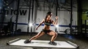 A woman in athletic workout clothes stretching on a yoga mat in a gym, with gym equipment and mirrors in the background.