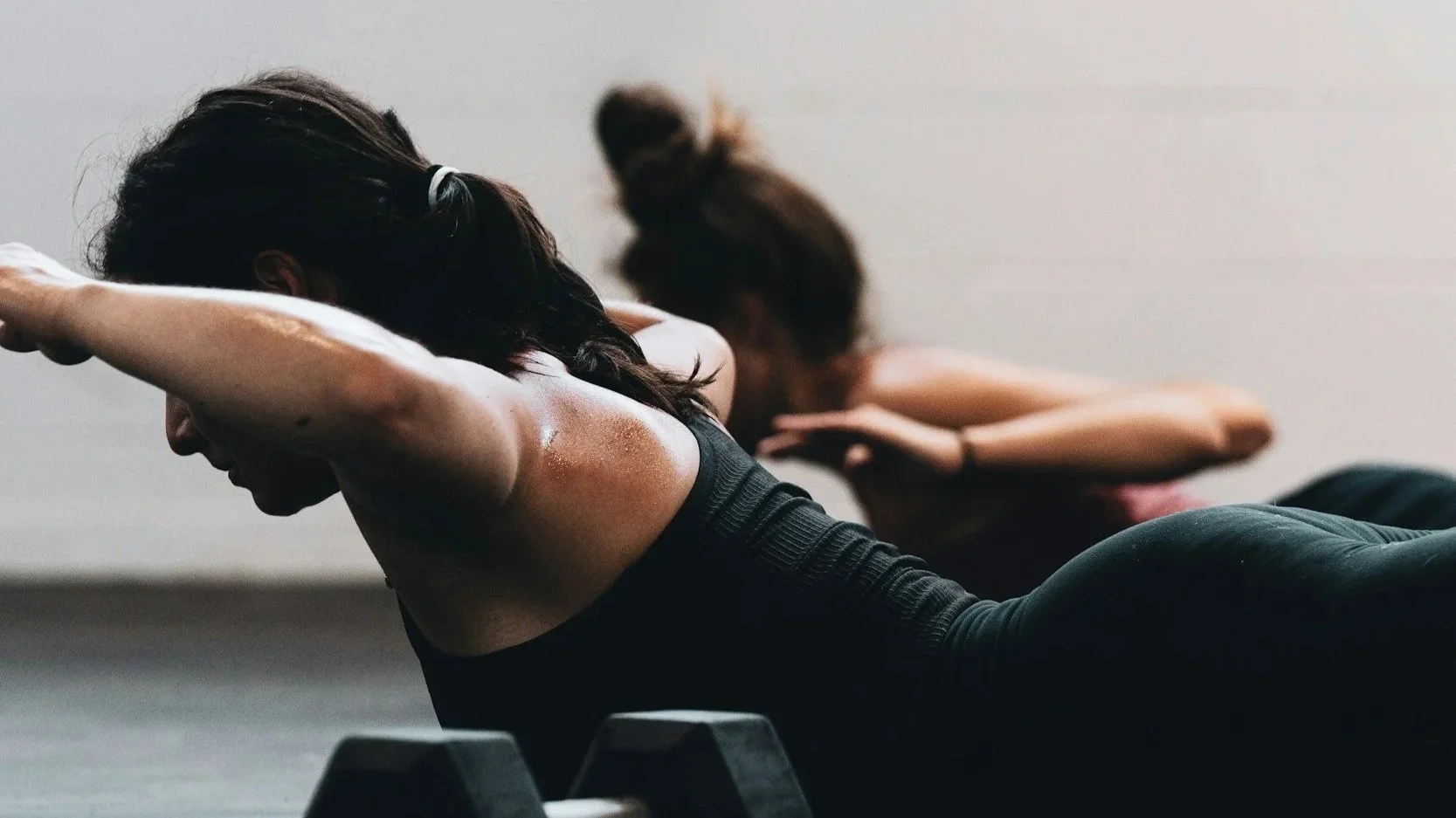 Two women doing core exercises on yoga mats in a fitness class, with a close-up of the woman in front with dark hair and black workout clothes.