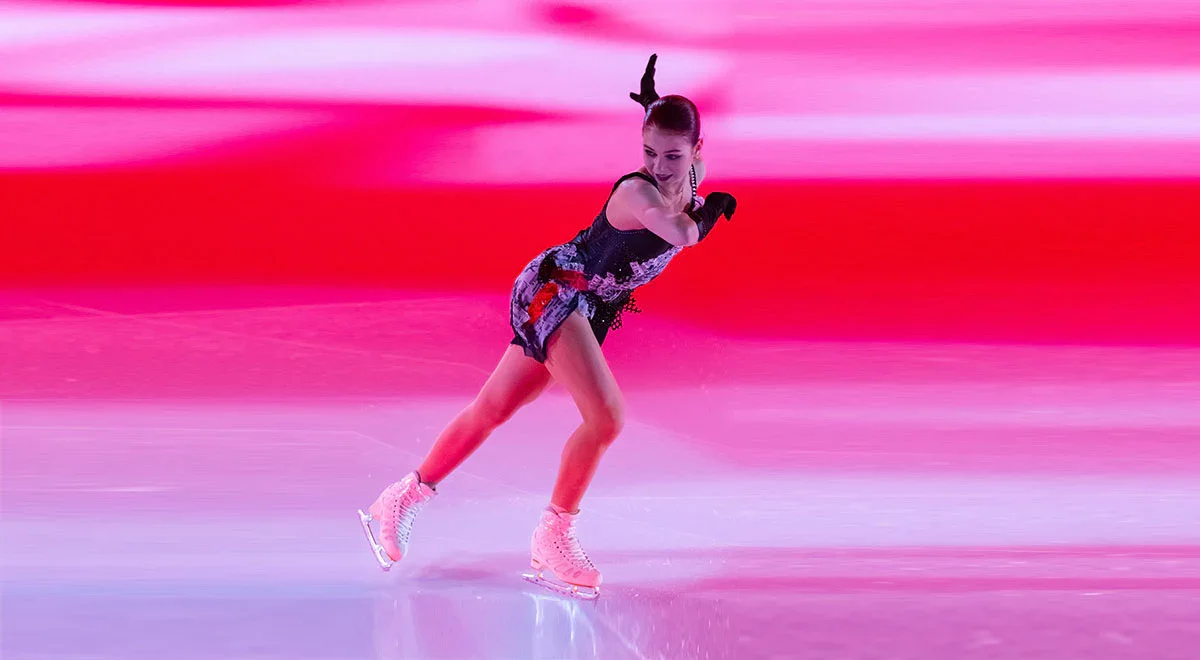 Figure skater wearing a black and purple costume gliding on ice rink with a pink and red background.
