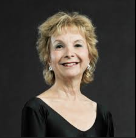 Portrait of an elderly woman with short, wavy blonde hair, smiling, wearing a black top against a dark background.