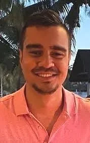 A man smiling outdoors with palm trees and a building in the background.