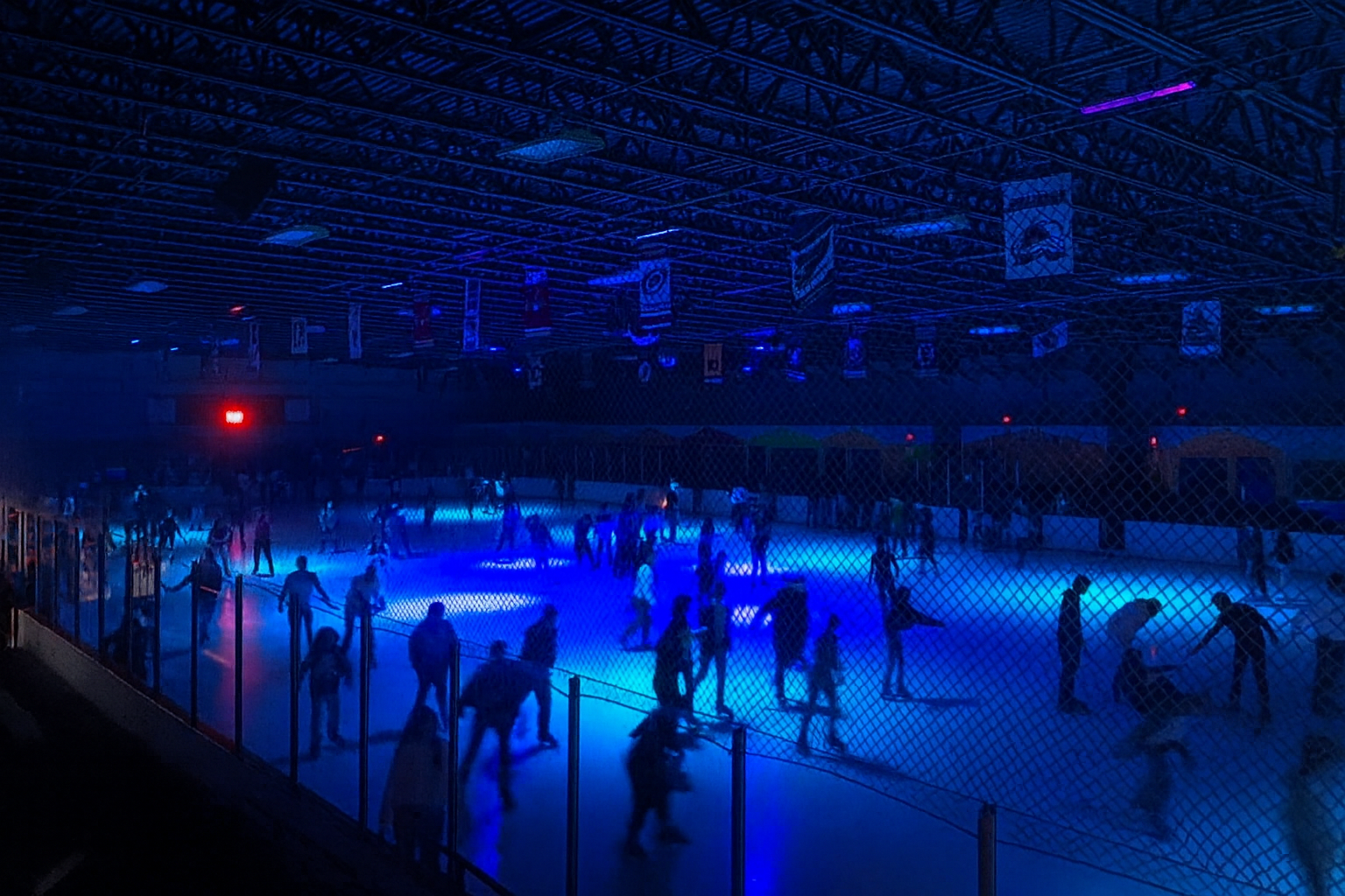 Indoor ice skating rink with blue lighting, people skating and sitting on the ice, fencing around the rink, banners hanging from the ceiling, and dark surroundings.