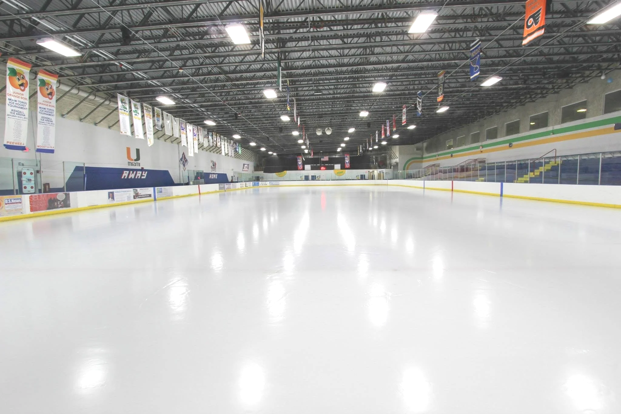 Empty indoor ice skating rink with white ice surface, surrounded by barriers and seating areas, inside a large sports arena with banners hanging from the ceiling.