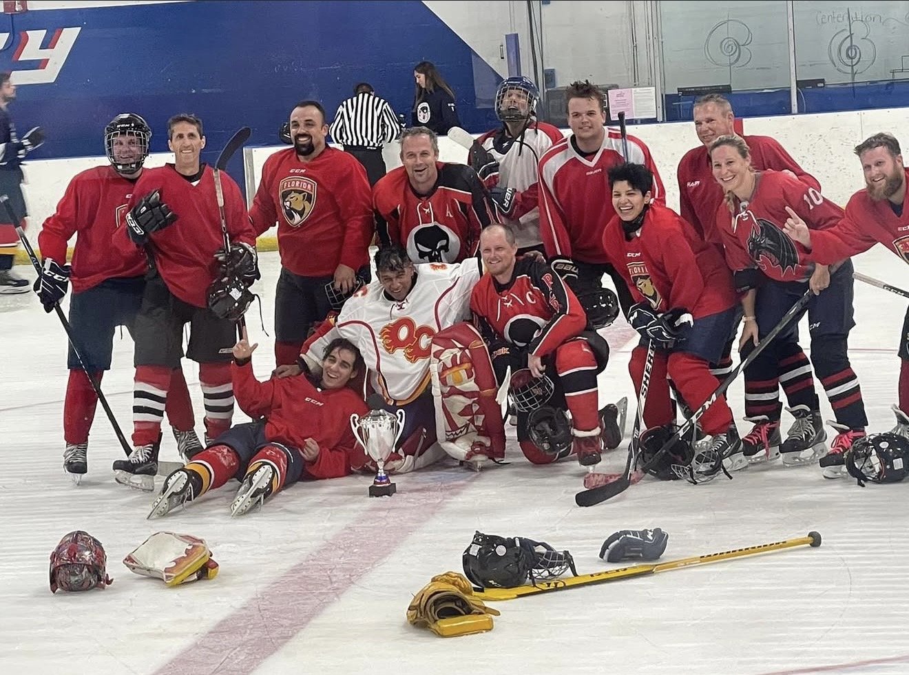 Group of ice hockey players in red and white jerseys celebrating on the ice rink after a game, with some players sitting or lying on the ice, and a trophy in front.