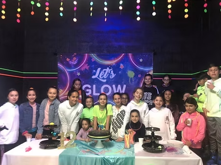 Group of children and teenagers celebrating a birthday party with a decorated table, birthday cake, and a 'Let's Glow' backdrop.