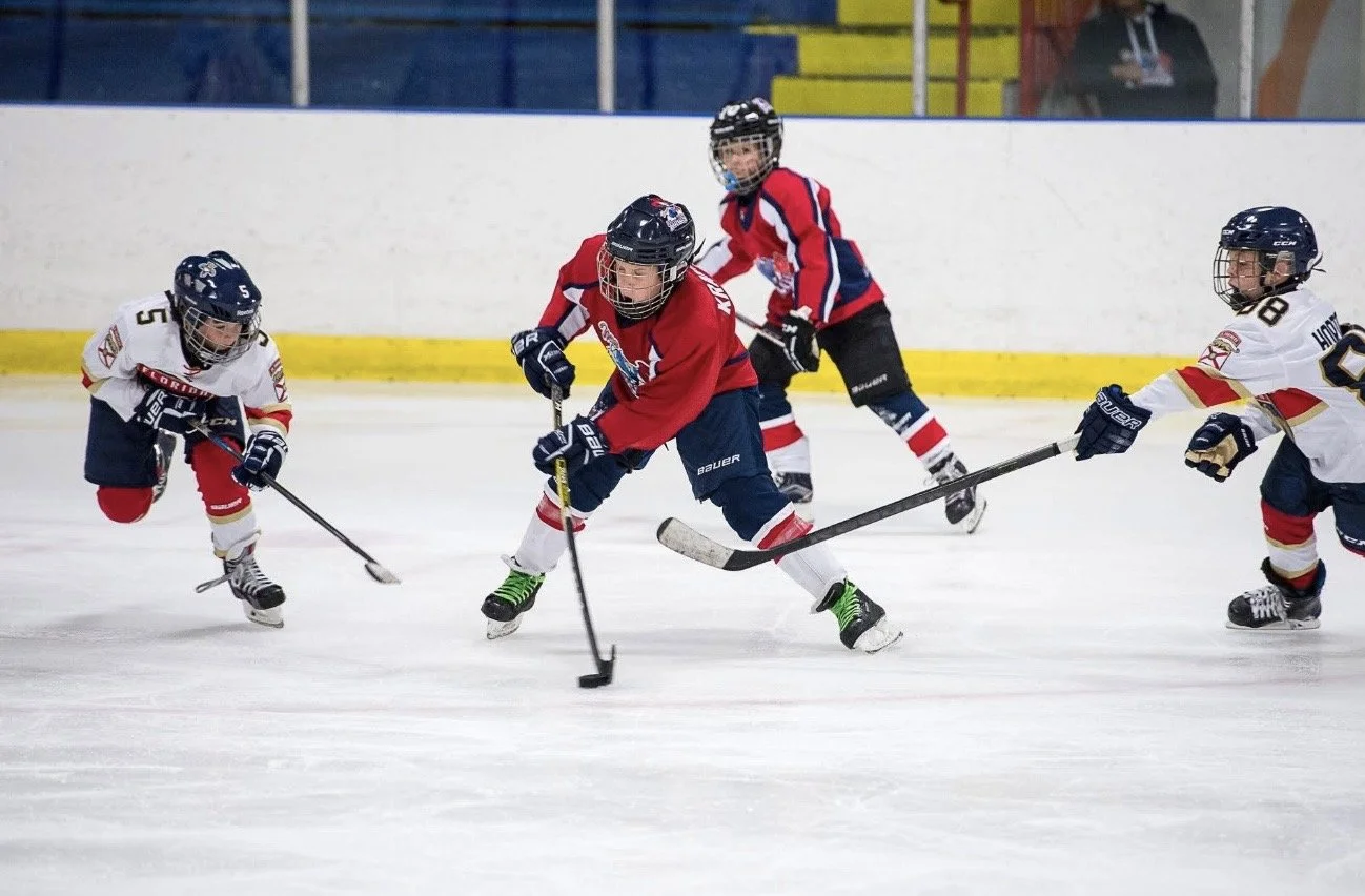 Young children playing hockey on the ice rink, wearing helmets and uniforms, competing for the puck.