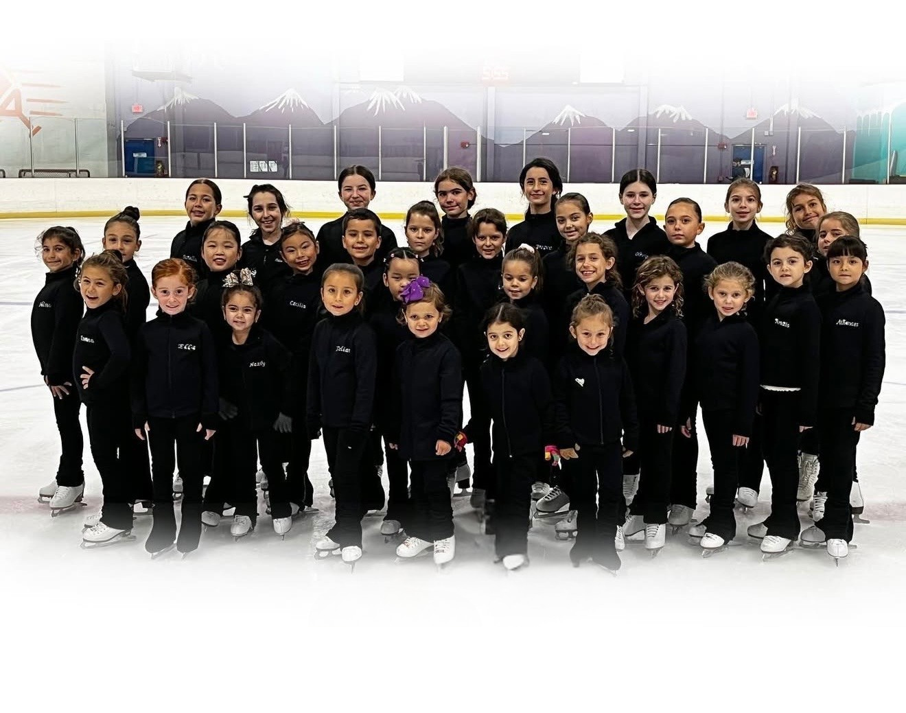 Group of young female figure skaters in black uniforms posing on ice rink.