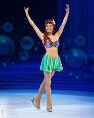 A person on ice skates wearing a purple and green costume, holding arms up in a pose, with a blue curtain backdrop.