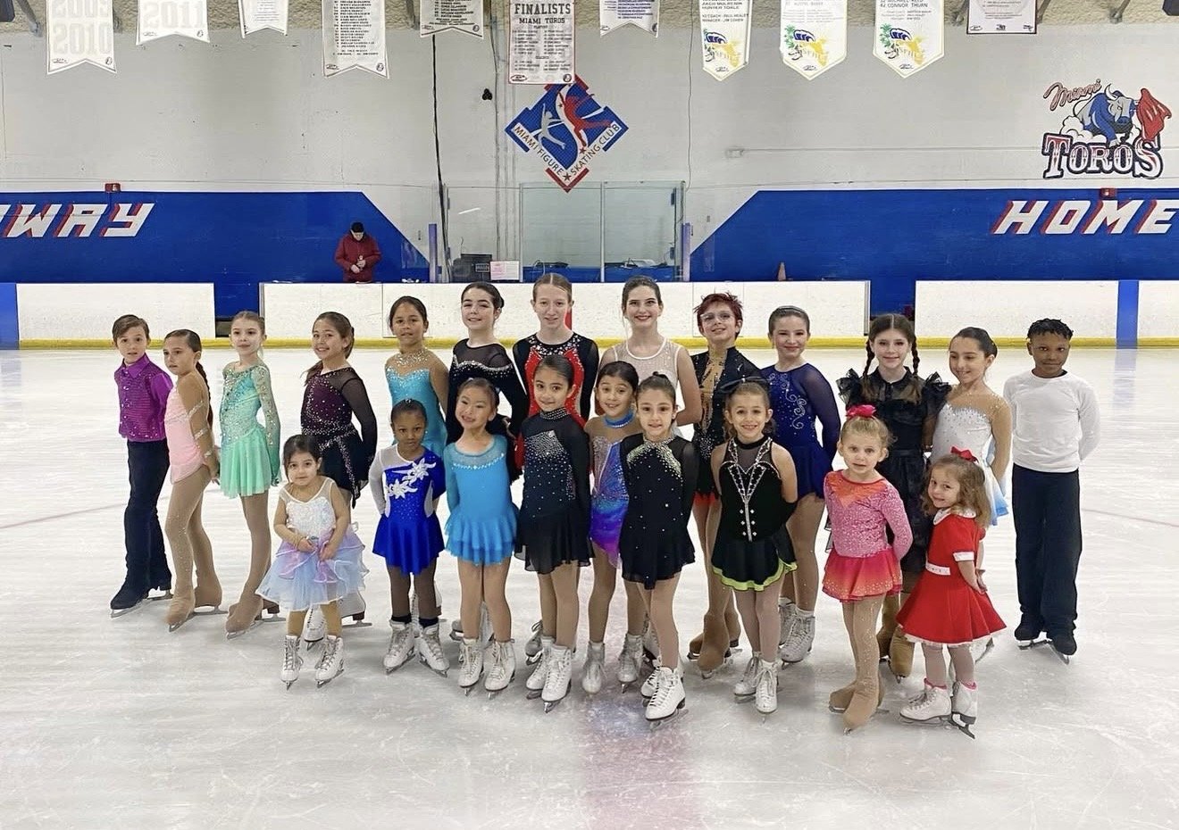 Group of young girls in colorful figure skating dresses and skates posing on ice rink, with coaches or staff in the background.