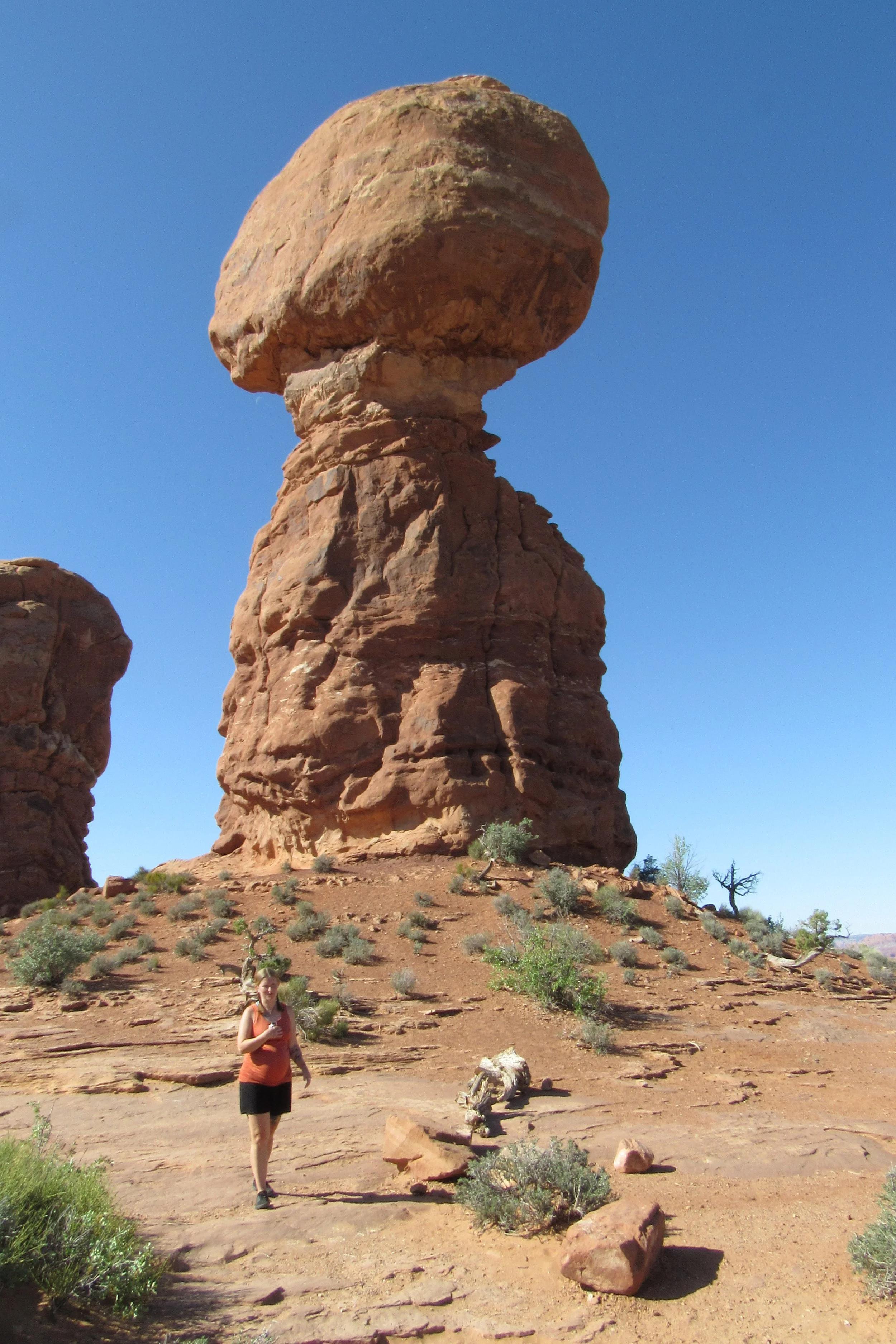 A person walking on a dirt trail in a desert landscape with large, uniquely shaped red rock formations, including a tall rock balanced with a massive boulder on top, under a clear blue sky.