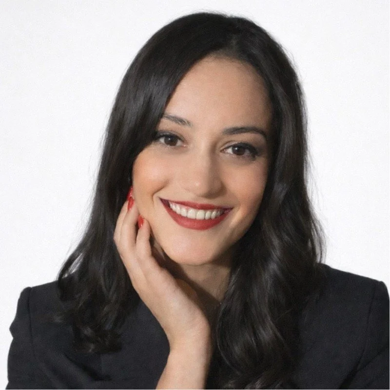 A smiling woman with long dark hair, wearing a black blazer, showing her teeth and with her hand touching her face, against a white background.