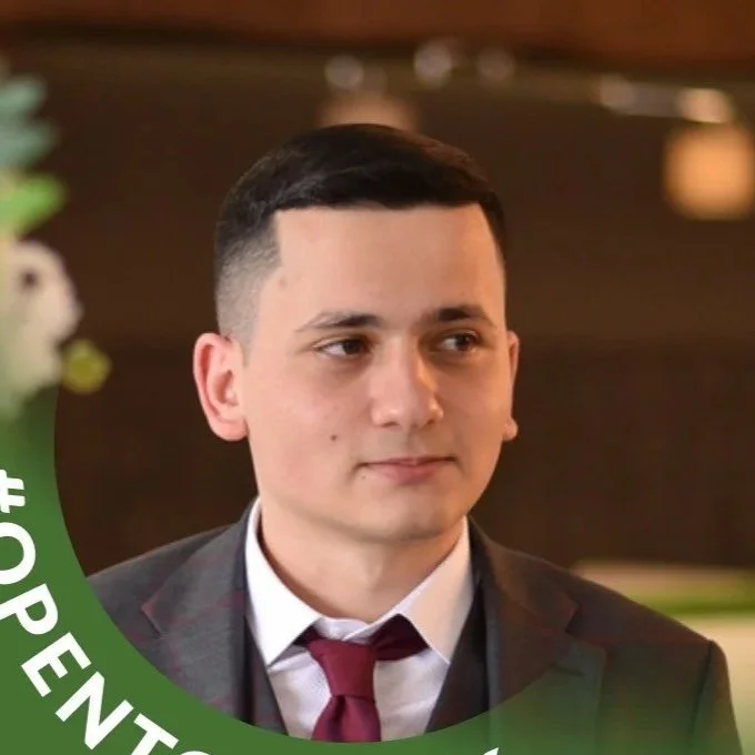 A young man in a suit and maroon tie, with short dark hair, looking slightly to the side, in a formal setting.