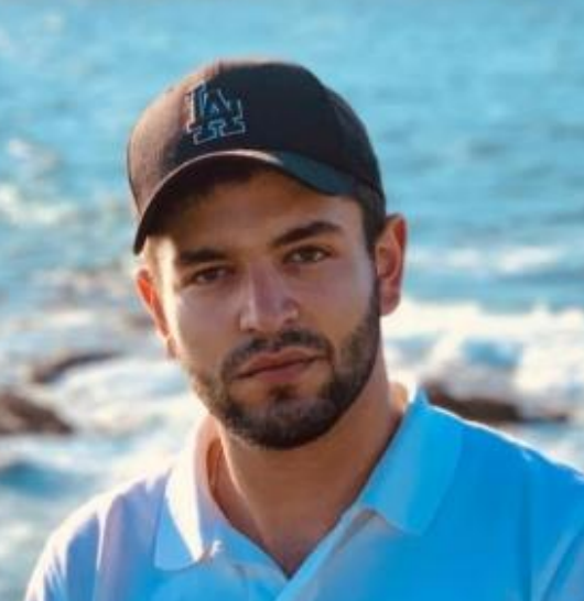 A man with dark hair and a beard wearing a black baseball cap and a light blue collared shirt, standing outdoors with a blurred ocean and rocks in the background.
