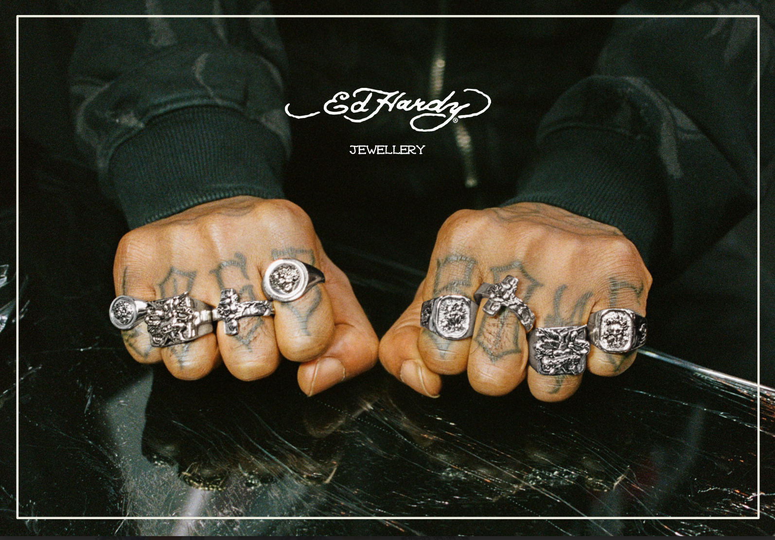Hands with tattoos wearing silver rings, displayed on a reflective black surface with 'Ed Hardy Jewelry' text overlay.