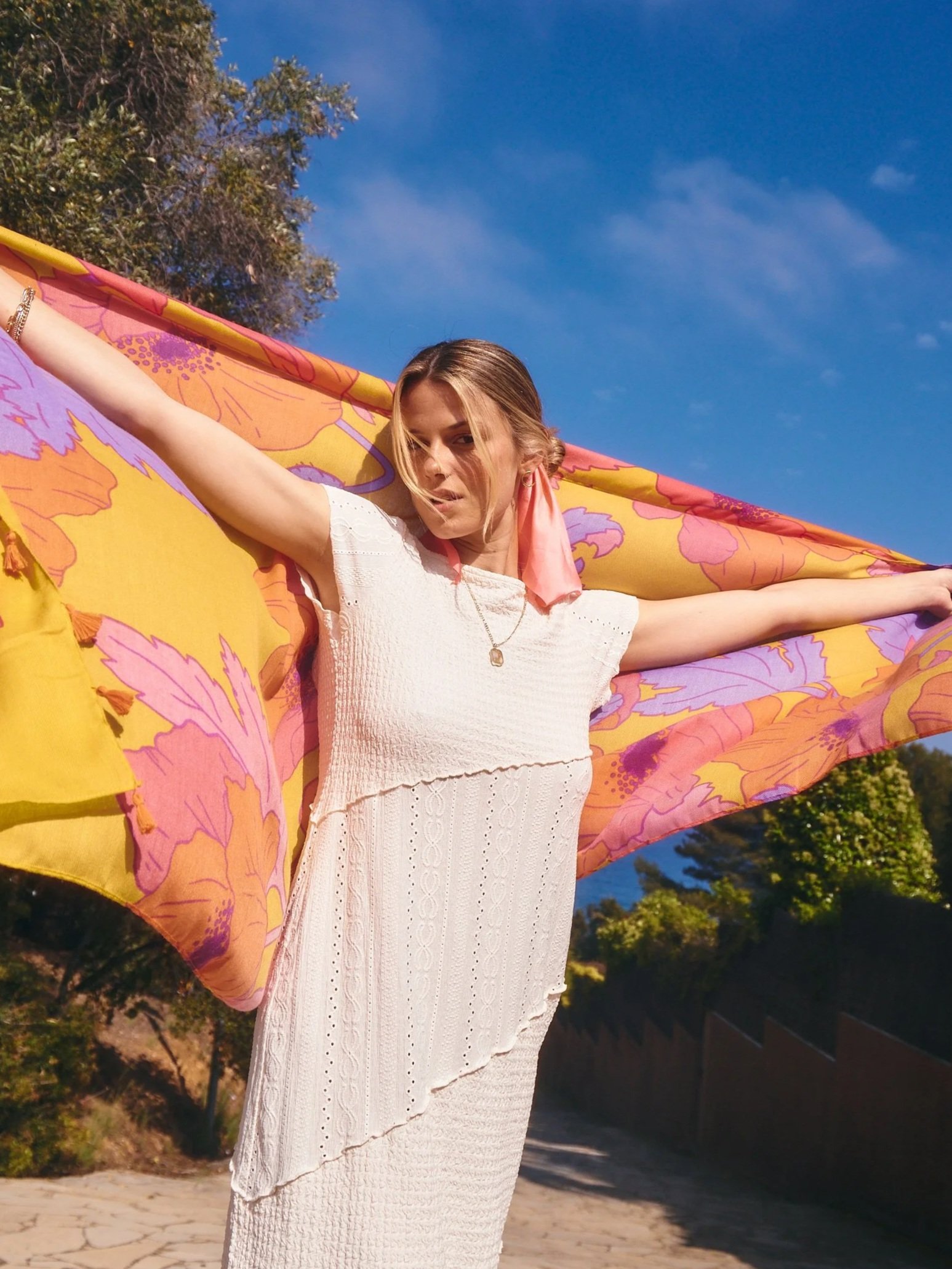 A woman standing outdoors under a clear blue sky, holding a colorful floral fabric or scarf outward with her arms extended. She is wearing a white dress with textured fabric and a pink scarf in her hair.