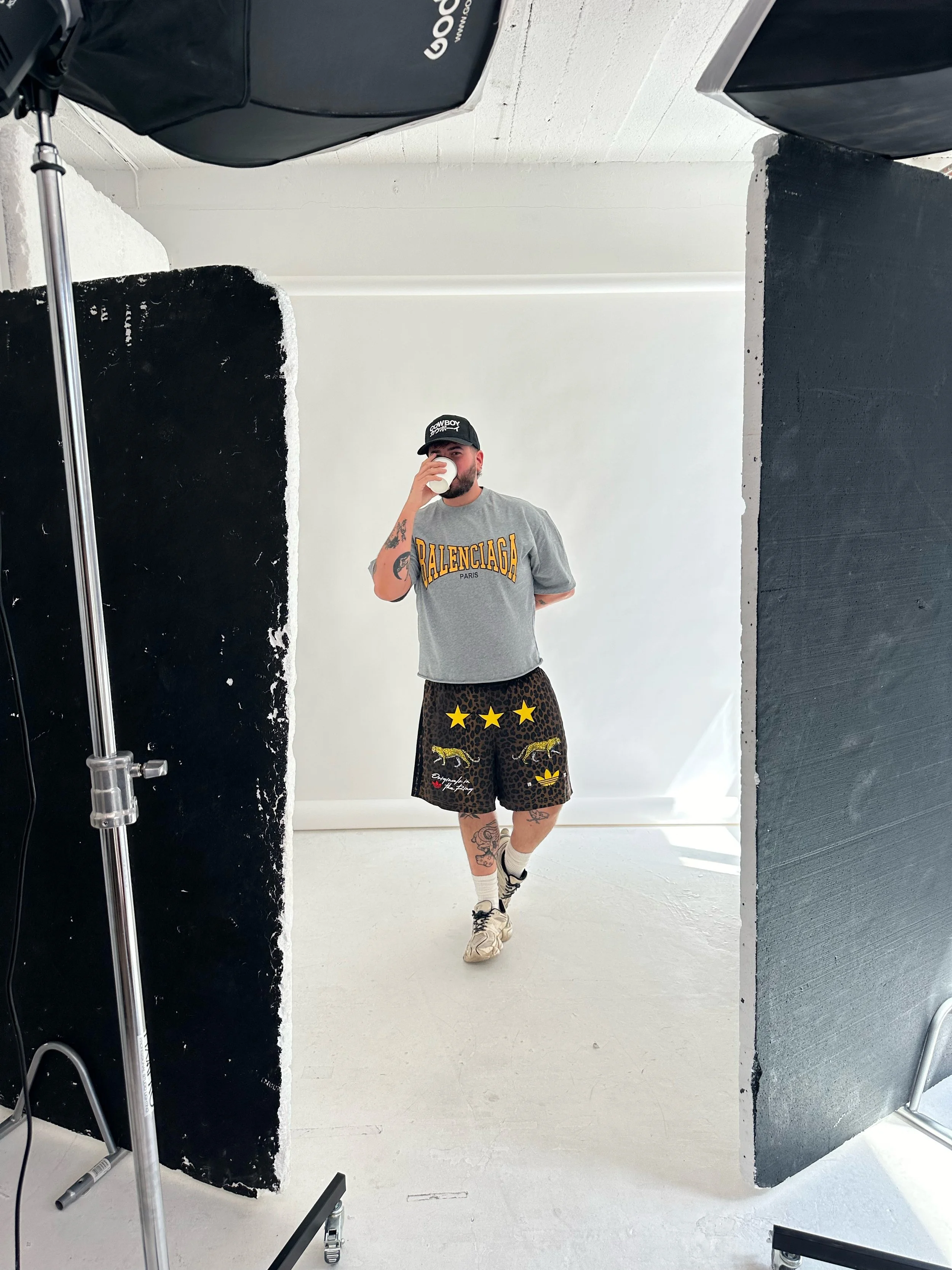 Person taking a mirror selfie in a photography studio, wearing a black cap, gray T-shirt, leopard-print shorts with star and animal applique, athletic shoes, and socks, holding a coffee cup.