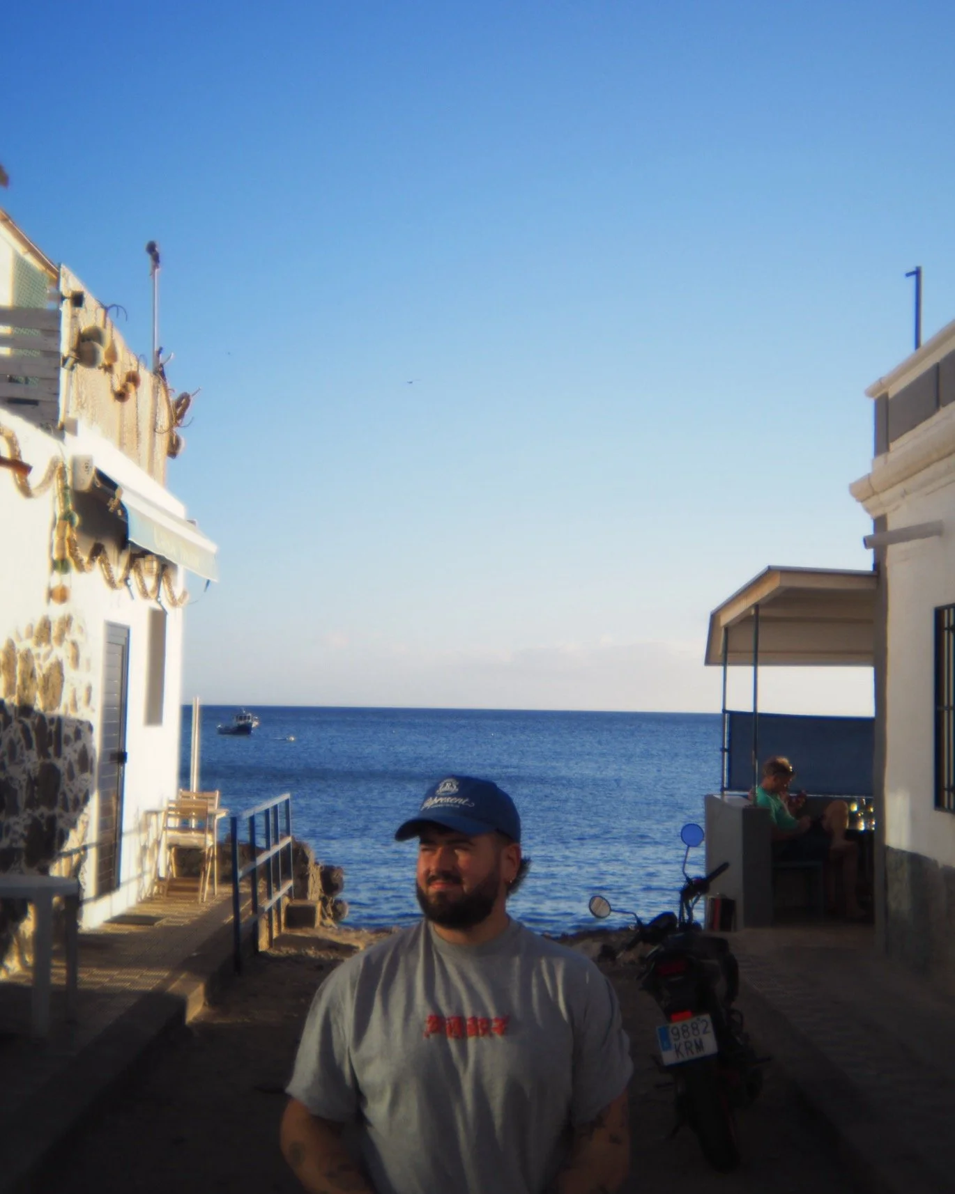 A man with a beard wearing a cap and gray T-shirt standing on a narrow street near the ocean, with a boat in the water and a person sitting in a shaded outdoor area.