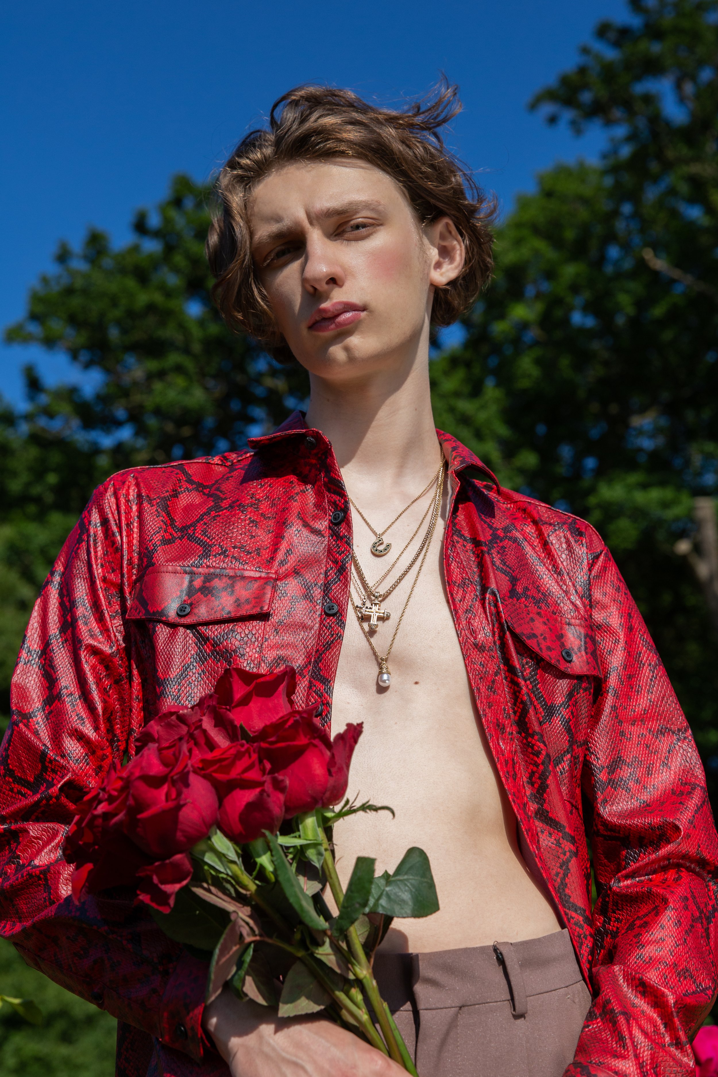 A young person with short, wavy brown hair holding a bouquet of red roses, wearing a red snakeskin-patterned shirt open at the front, tan pants, and layered gold necklaces, outdoors on a sunny day with green trees in the background.