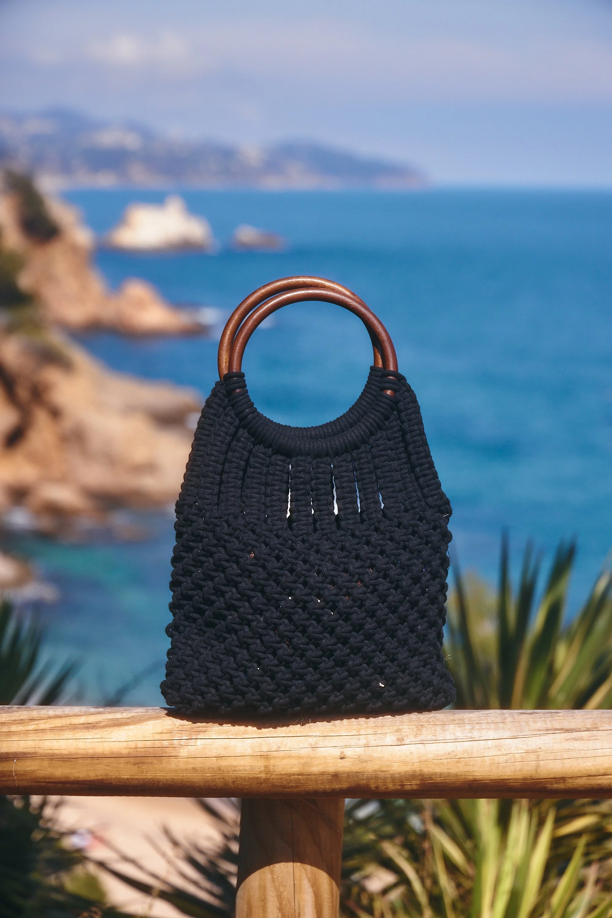 A black woven bag with wooden handles sits on a wooden railing, with a scenic ocean view and rocky cliffs in the background.