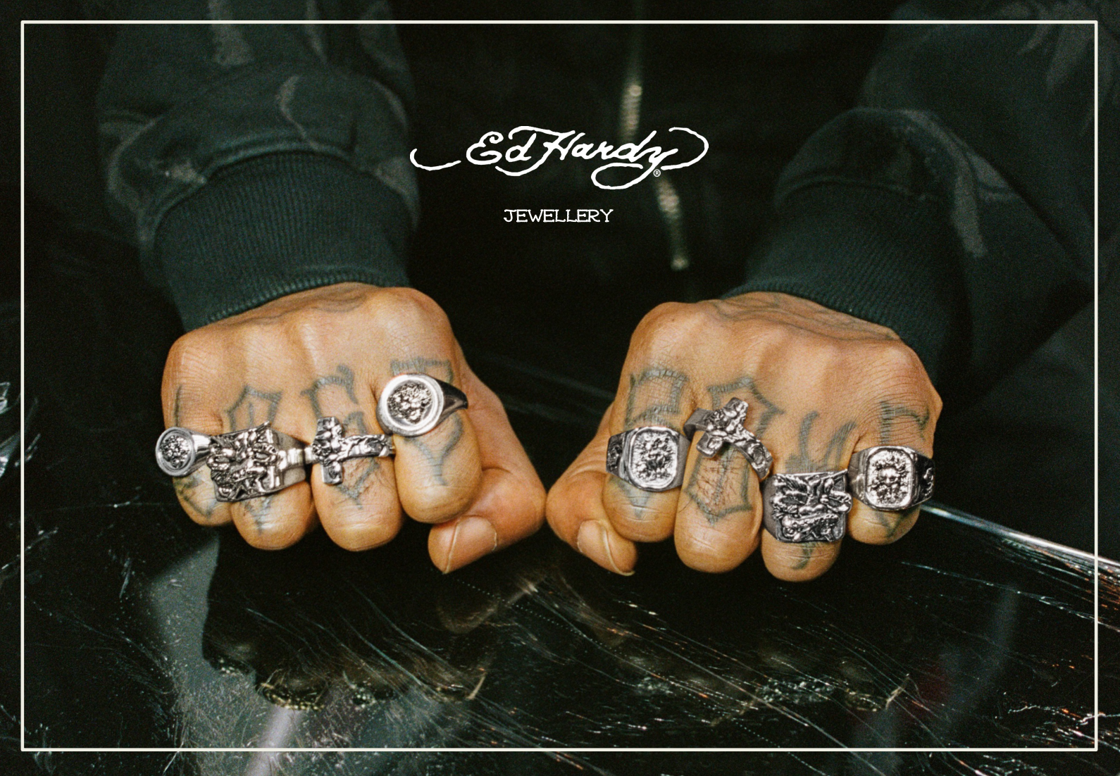 Close-up of a person's hands with jewelry and tattoos, wearing rings with lions and other designs, on a reflective black surface, with Ed Hardy jewelry logo at the top.