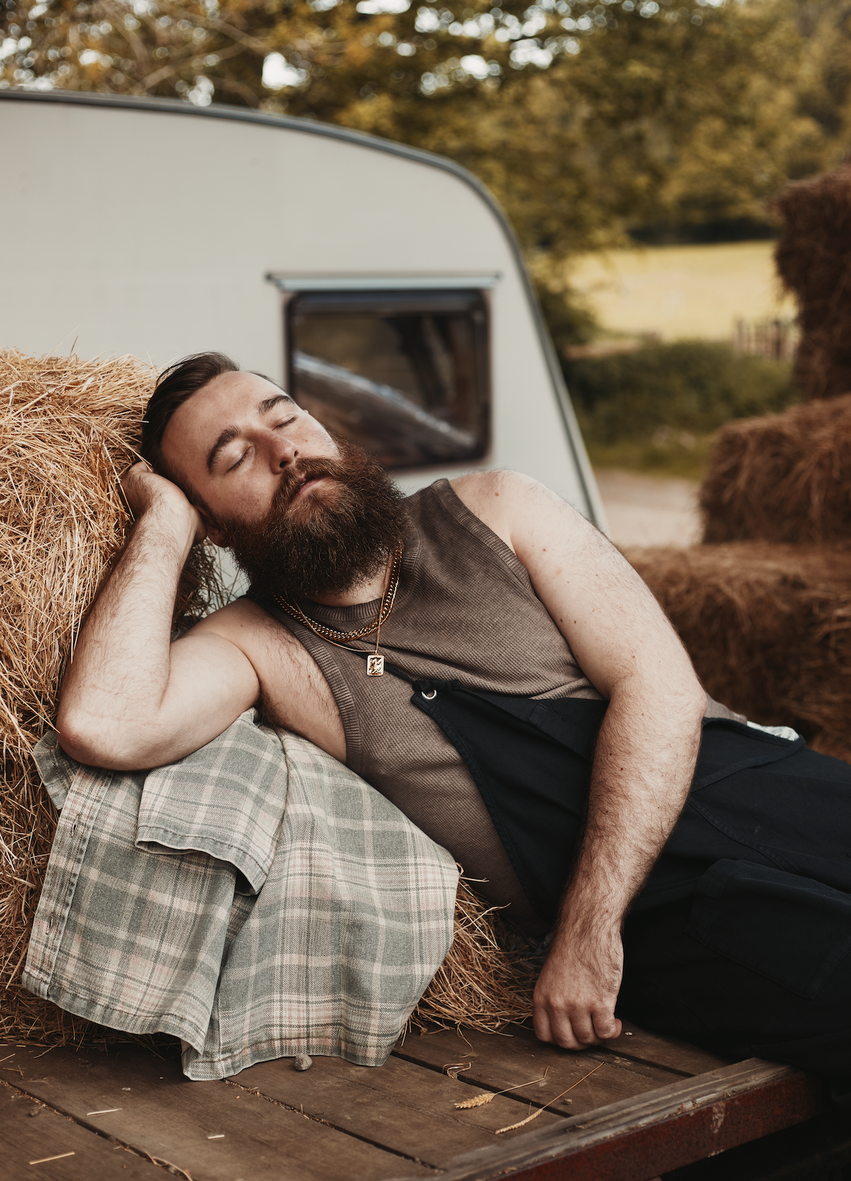 A man with a beard and tattoos sleeping on a hay bale, leaning against a plaid shirt and wooden surface, outdoors with trees and a travel trailer in the background.