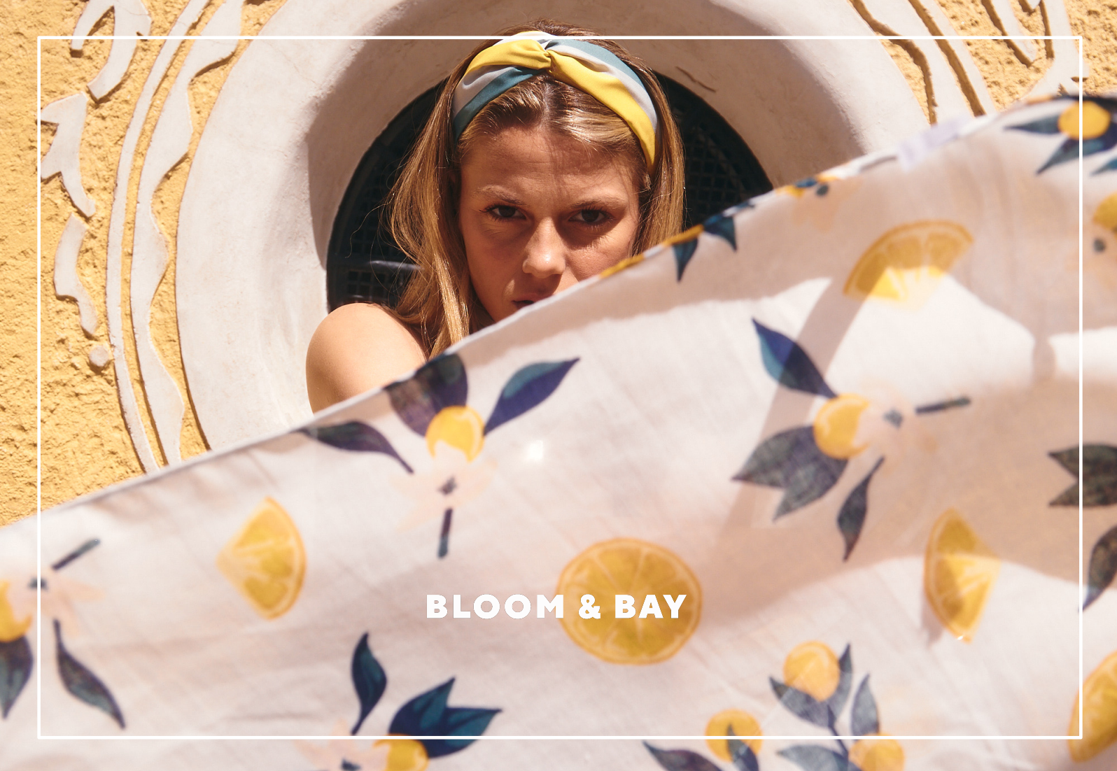 A woman with blonde hair wearing a yellow and blue headband, looking intensely at the camera while holding a white cloth with lemon and leaf patterns in front of her face. She is standing in front of a yellow wall with decorative white and gold accents, and there is a white arched window or opening behind her. The lower part of the image contains the text 'BLOOM & BAY' in white letters.