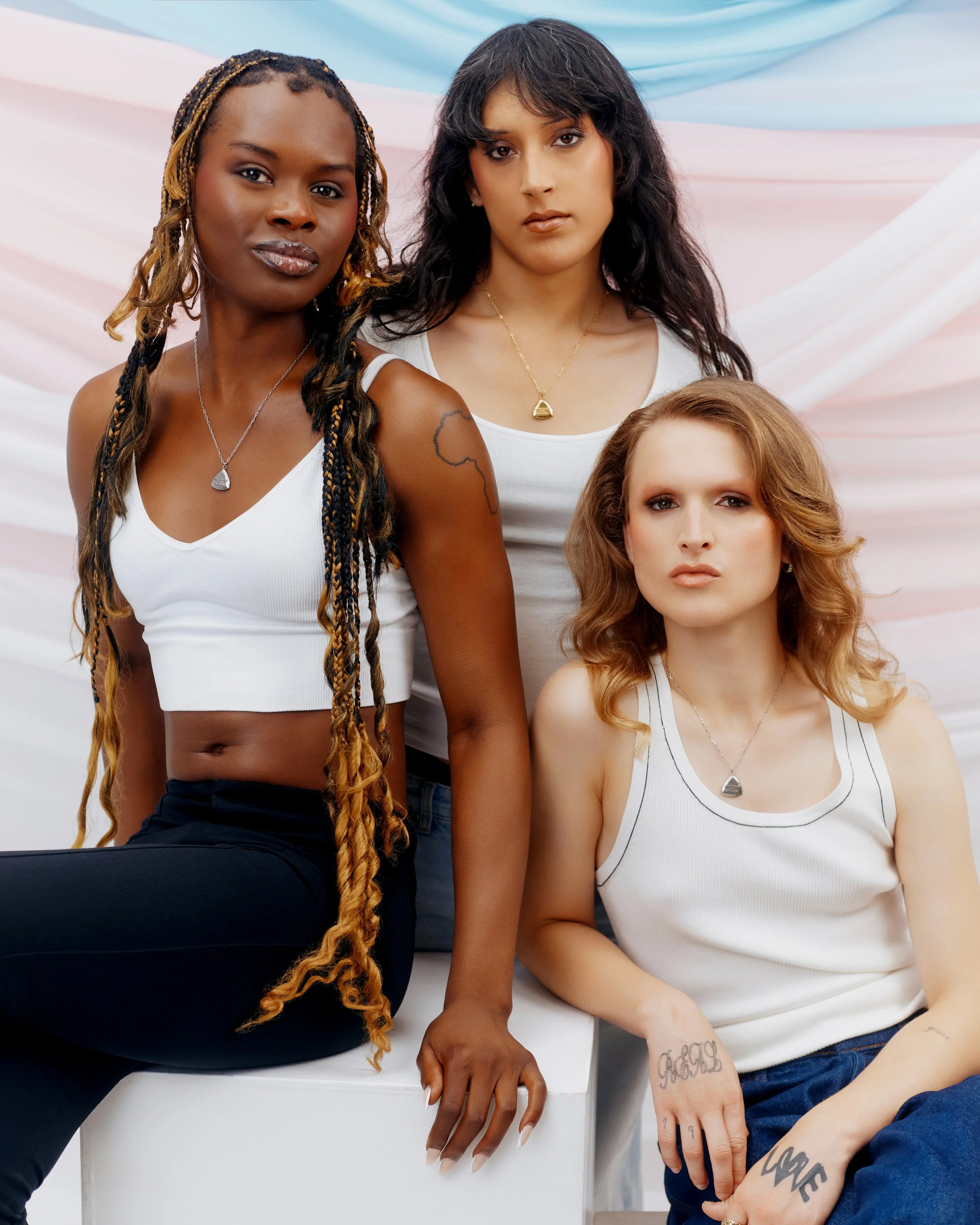 Three women with different hair styles and skin tones posing together against a pastel pink and blue backdrop. One woman is sitting on a white block, and the other two women are standing behind her.