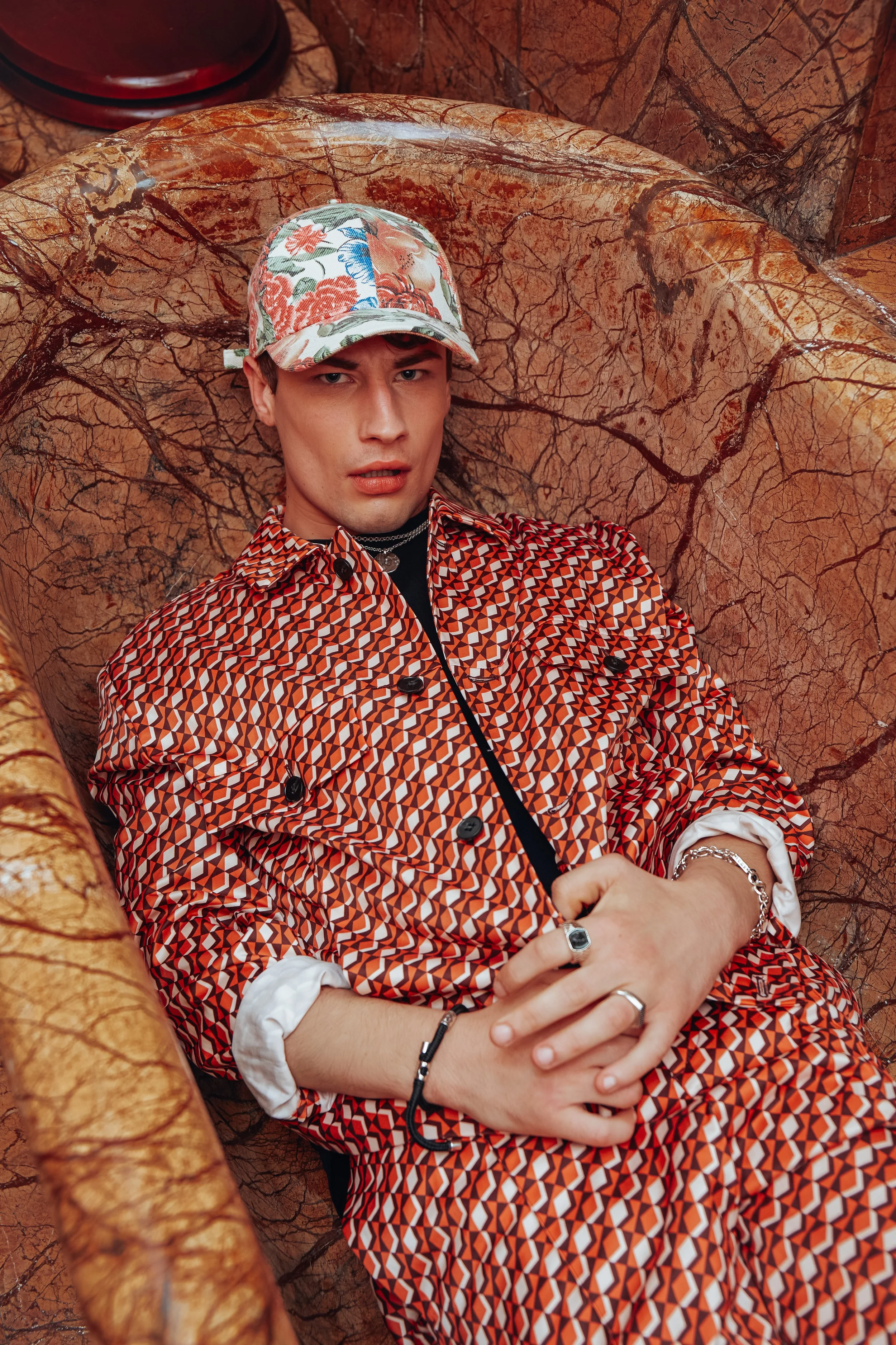 A young man wearing a colorful floral patterned cap and a red geometric patterned coat lying on a marble bench with a matching marble background.
