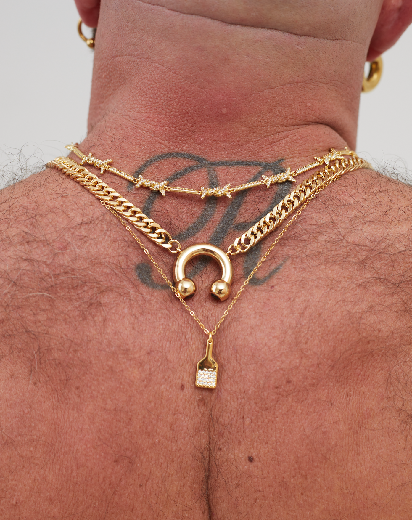 Close-up of a person's neck with multiple gold necklaces, including a chain with a lock and charms, a choker with small charms, and a pendant with a diamond-studded charm.
