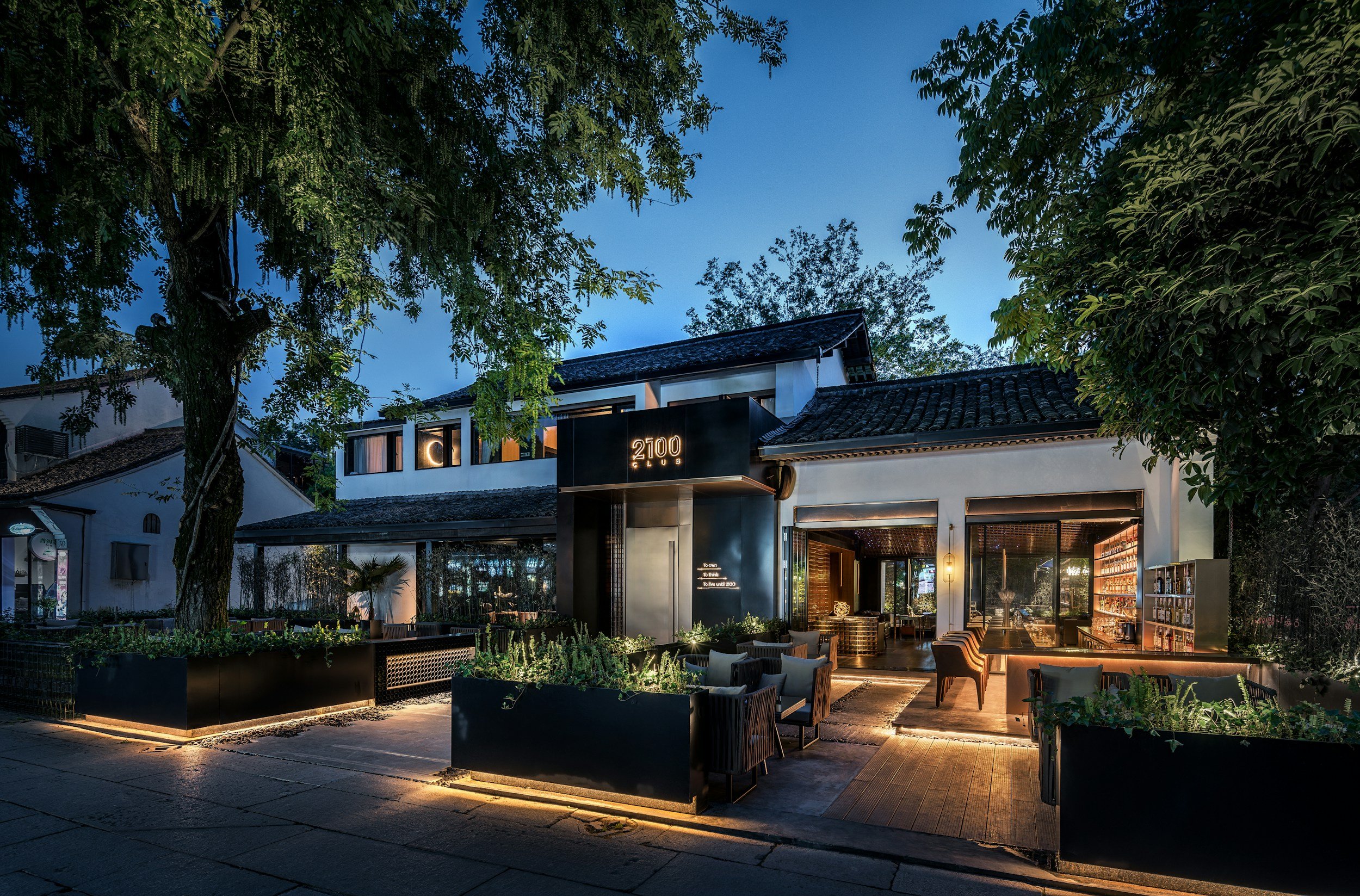 A stylish restaurant or café at dusk with outdoor seating, illuminated by warm lighting, surrounded by greenery and tall trees.