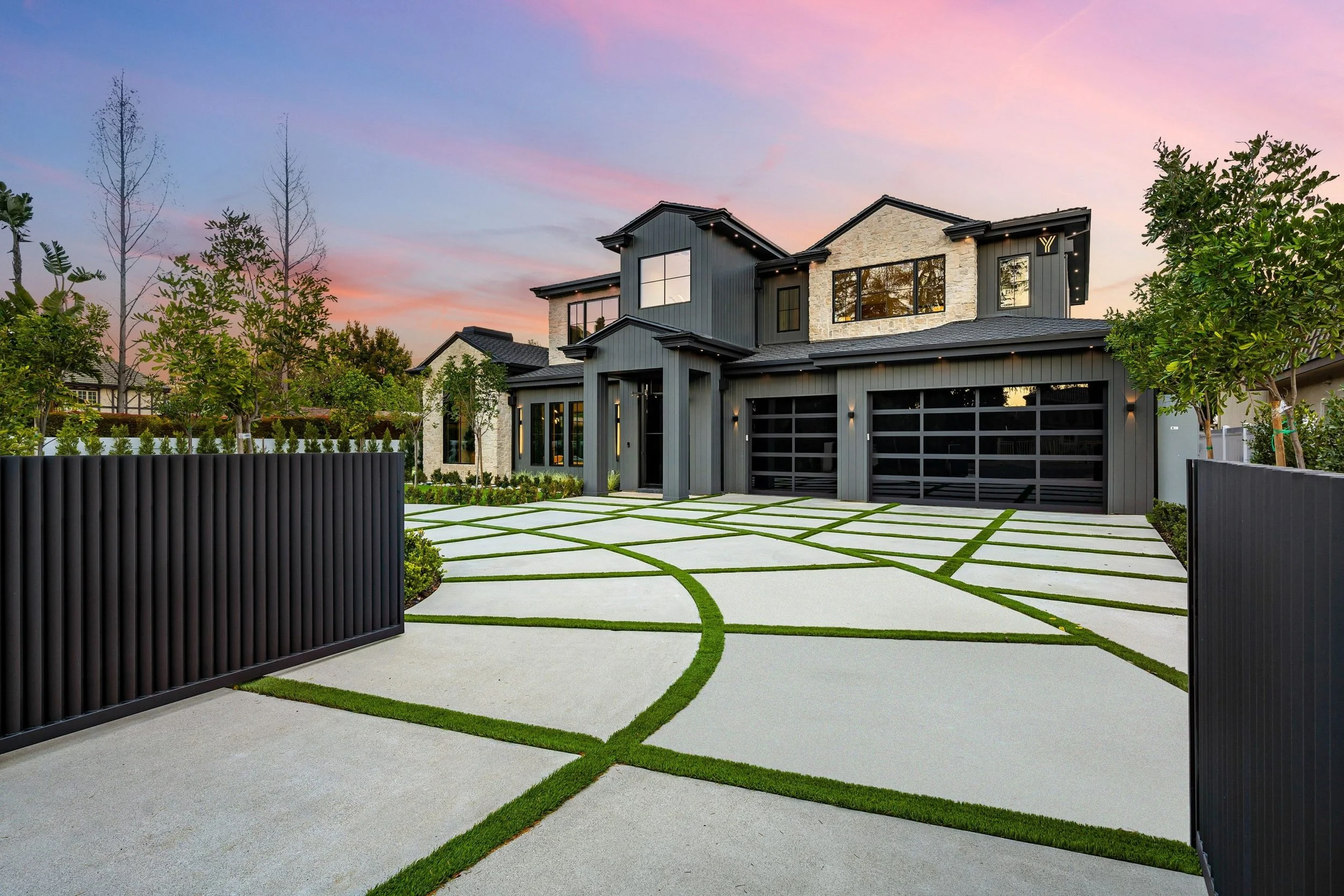 Modern two-story house with dark exterior, large windows, and a garage with black doors, surrounded by a landscaped yard with patterned concrete driveway and green grass accents, under a pink and purple sunset sky.
