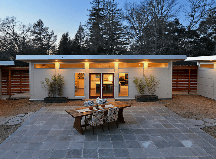 Modern house with a patio featuring a wooden dining table and six chairs with floral upholstery, two planters with shrubs, and large glass doors leading inside, with a backdrop of trees and a clear sky.
